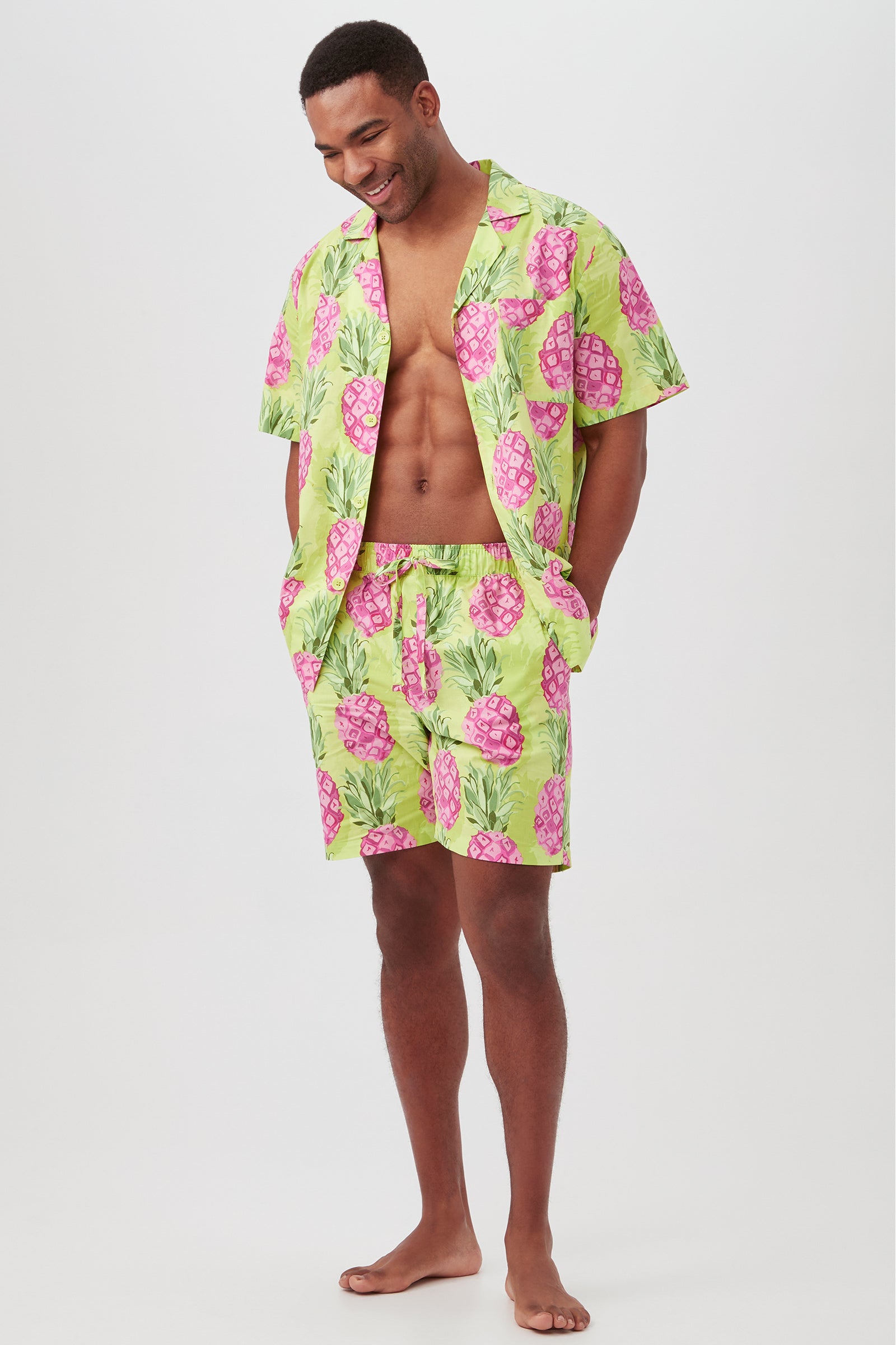 A man stands barefoot, smiling, in the Kiwi Pineapple Mens Short Sleeve Boxer Short Poplin PJ Set with a bold green and pink pineapple print. The open button-up shirt reveals his chest, and he rests his hands in his pockets.