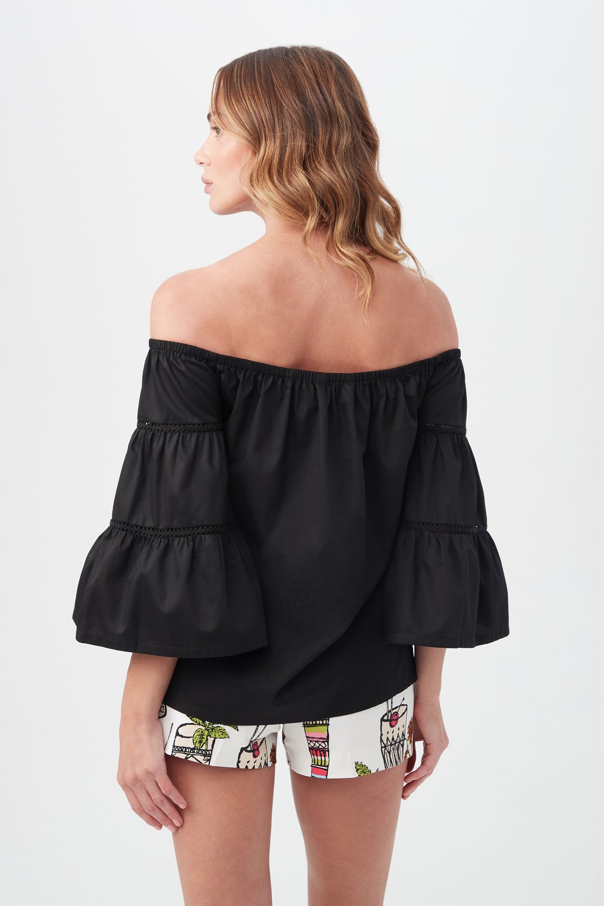 A woman with wavy, shoulder-length hair models the Garnish Top—an off-the-shoulder black blouse with tiered sleeves—paired with colorful patterned shorts, shown from the back against a plain white background.