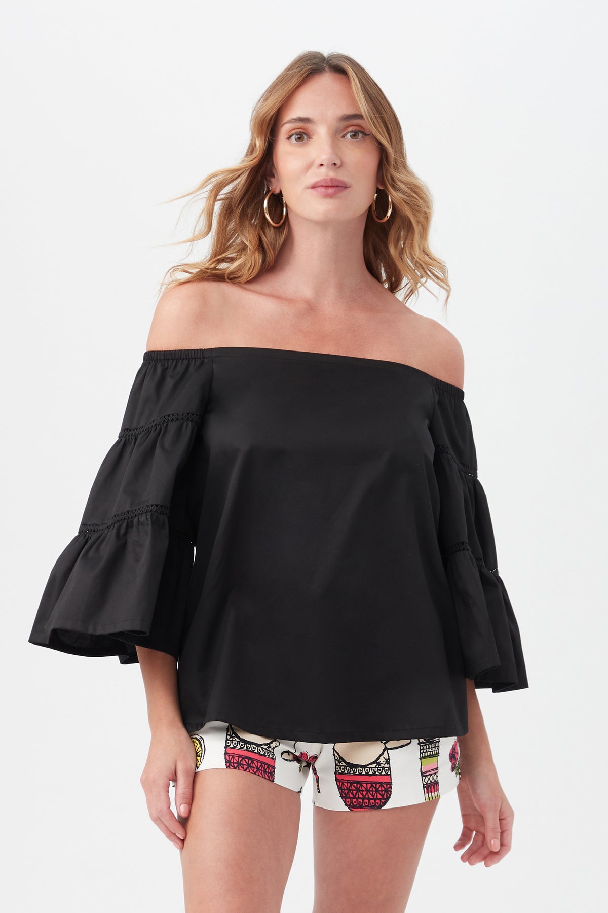 A woman with wavy light brown hair and hoop earrings models the black Garnish Top with flared sleeves, paired with patterned white shorts, standing against a plain white background.