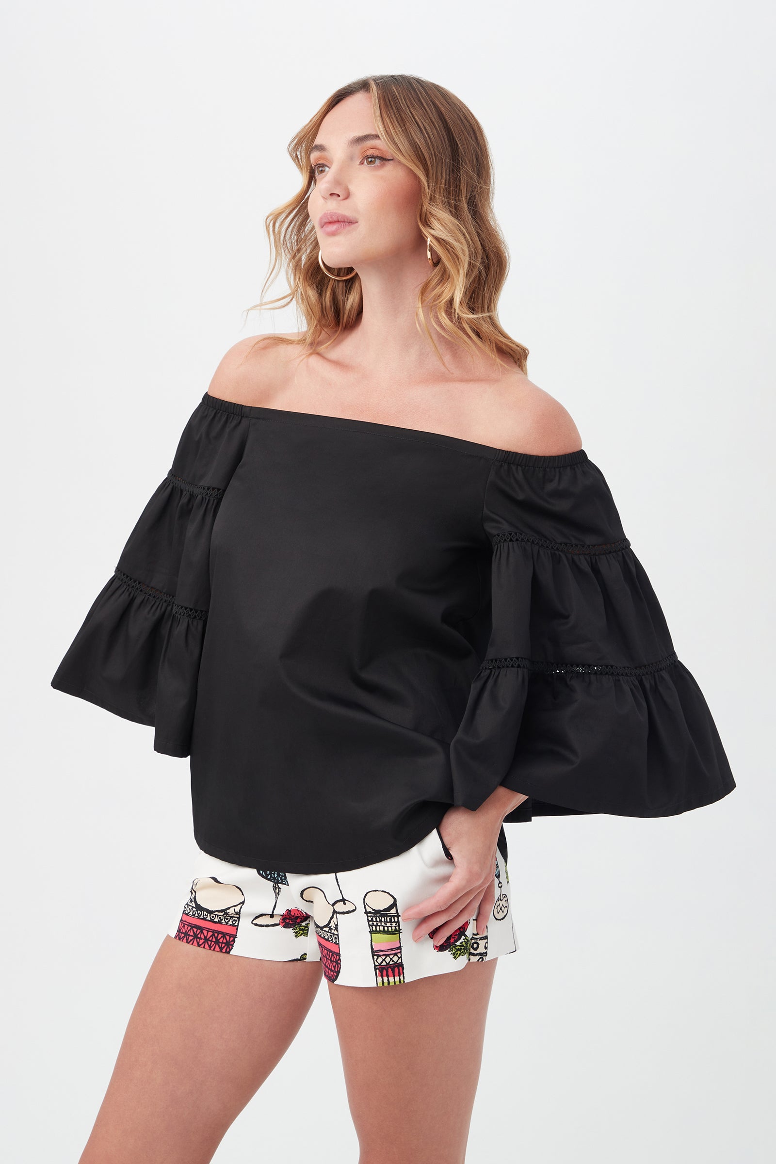 A woman wears the Garnish Top, an off-the-shoulder black blouse with wide, ruffled sleeves, paired with white shorts featuring colorful graphic designs, standing against a plain light background.