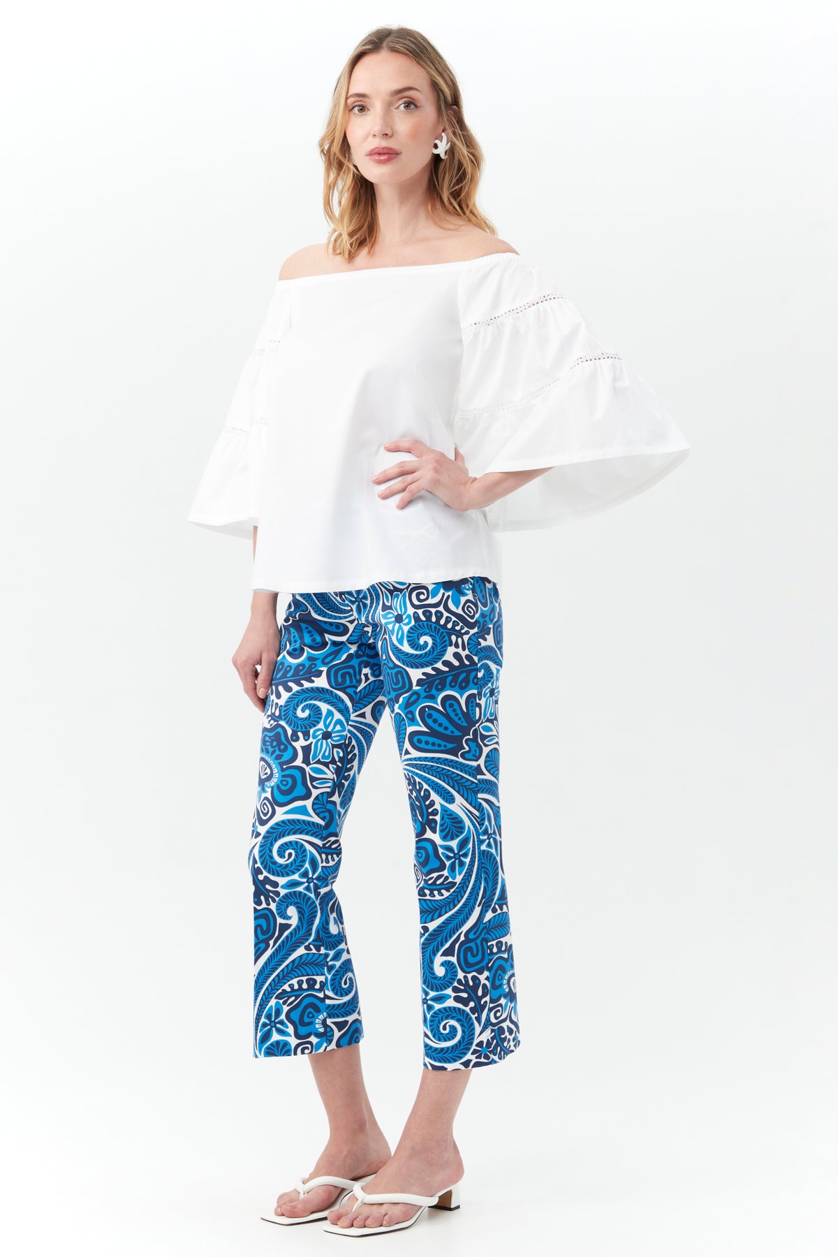A woman stands against a white background, wearing the Garnish Top—an off-the-shoulder white blouse with flared sleeves—paired with blue and white patterned pants, white sandals, and star-shaped earrings.