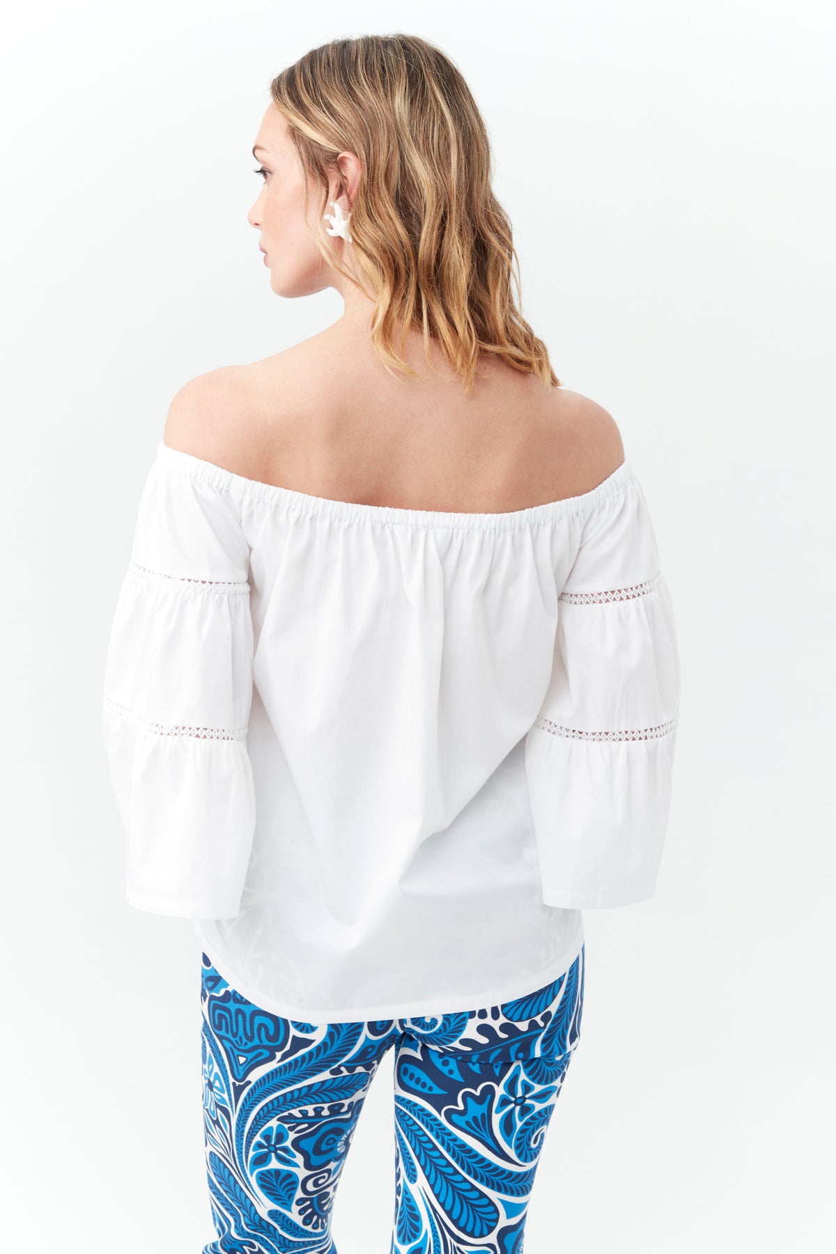 A woman with wavy blonde hair faces away, wearing the Garnish Top—an off-the-shoulder white blouse with long sleeves—and blue pants with a bold paisley pattern on a white background.