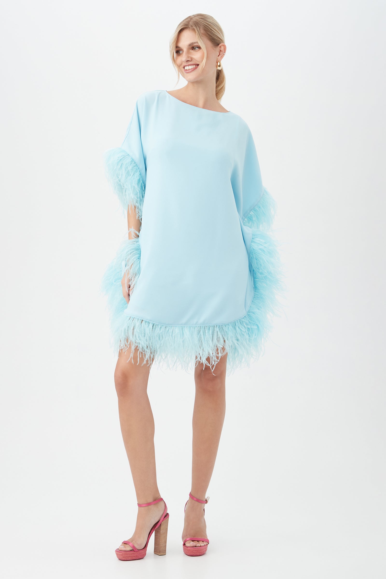 A woman smiles in the Mini Neena Boatneck Dress With Feather Trim, a light blue, loose-fitting style with feather accents on the sleeves and hem. She pairs it with pink high-heeled sandals, her blonde hair tied back against a plain white background.