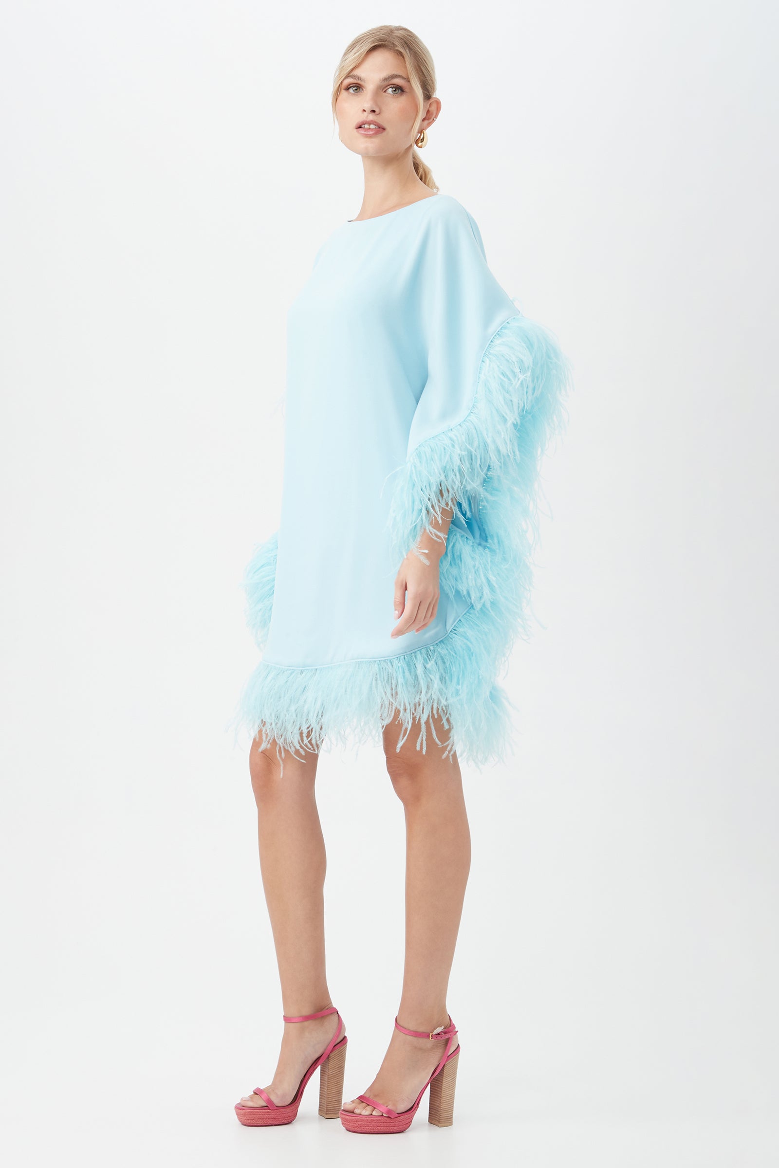 A woman stands against a white background, wearing the Mini Neena Boatneck Dress With Feather Trim in light blue, paired with pink platform heels. She gazes to the side, one hand slightly raised.