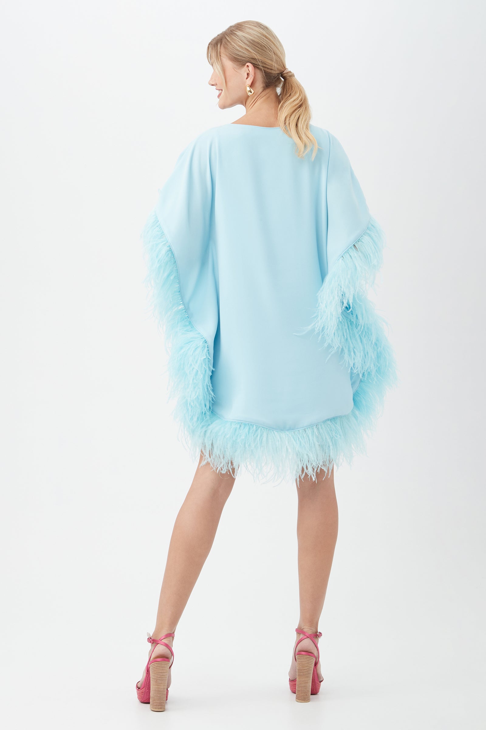 A woman faces away, wearing the Mini Neena Boatneck Dress With Feather Trim in light blue. Her blonde hair is in a low ponytail, and she pairs the dress with pink high heels and hoop earrings.