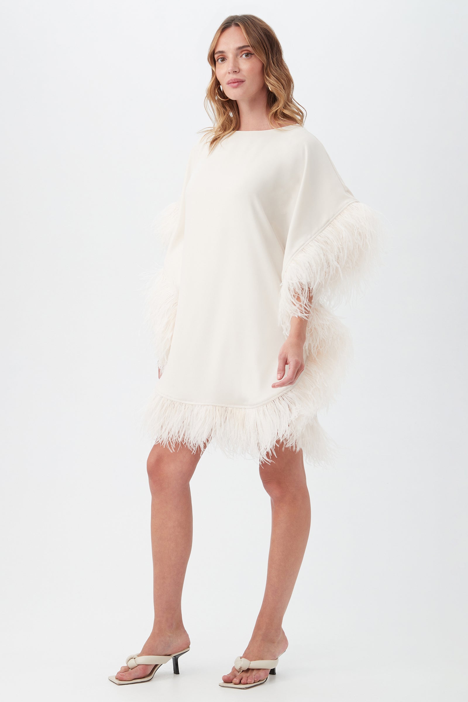 A woman poses against a plain white background wearing the Mini Neena Boatneck Dress With Feather Trim—an off-white, loose-fitting dress featuring feather details on the sleeves and hem, styled with wavy light brown hair and white heeled sandals.