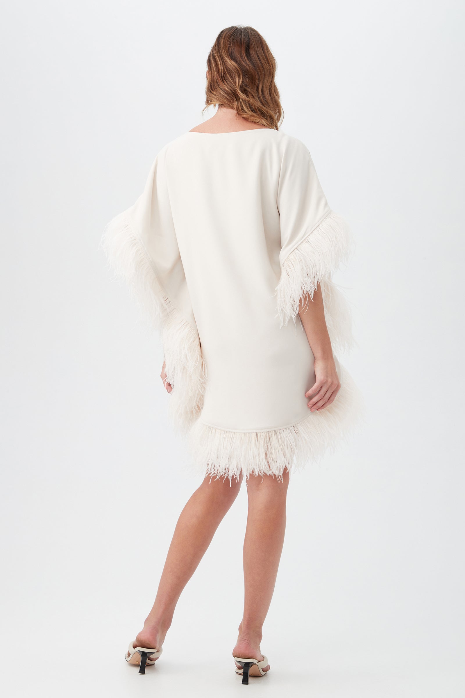 A woman with wavy brown hair faces away in the Mini Neena Boatneck Dress With Feather Trim, a loose cream knee-length dress featuring feather-trimmed sleeves and hem. She pairs it with open-toed heels, set against a plain white background.