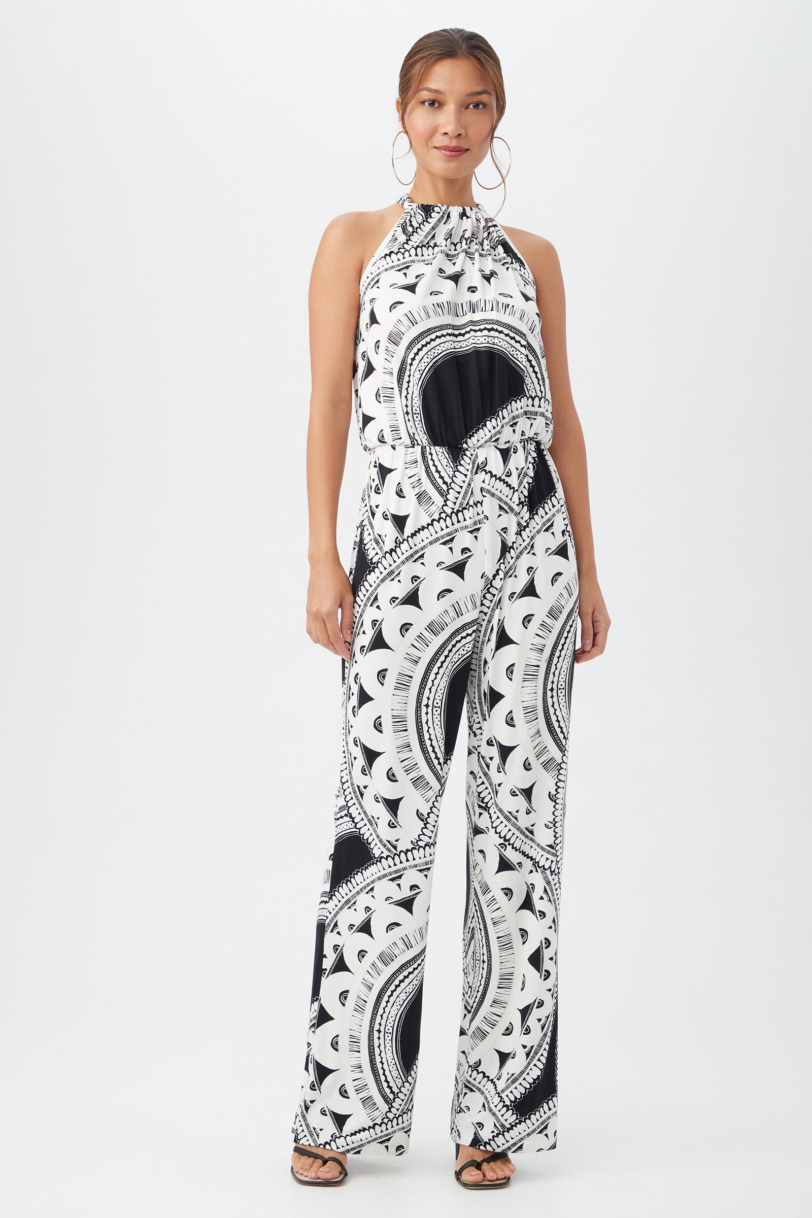 A woman poses against a white background wearing the Rancho Jumpsuit in multi, featuring a sleeveless design, high neck, wide-leg pants, and styled with hoop earrings and open-toe sandals.