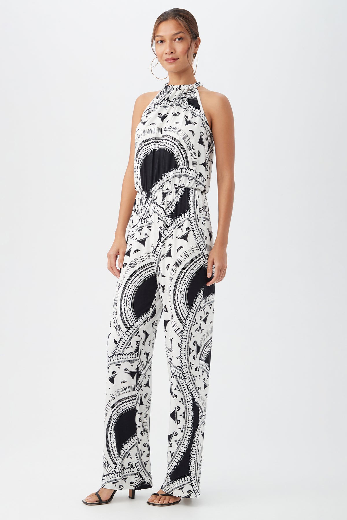 A woman poses against a plain white background in the Rancho Jumpsuit, a sleeveless, high-neck black and white patterned wide-leg style. She pairs it with black heeled sandals and has her hair tied back.