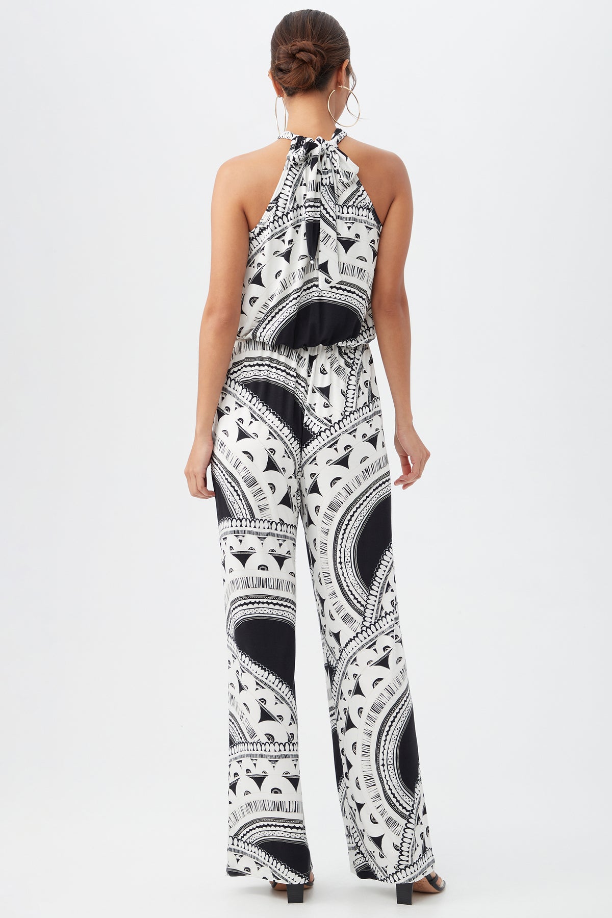 A woman stands against a white background, wearing the Rancho Jumpsuit—a sleeveless black and white patterned halter style with wide legs and an open back. She pairs it with hoop earrings, a low bun hairstyle, and black heels.