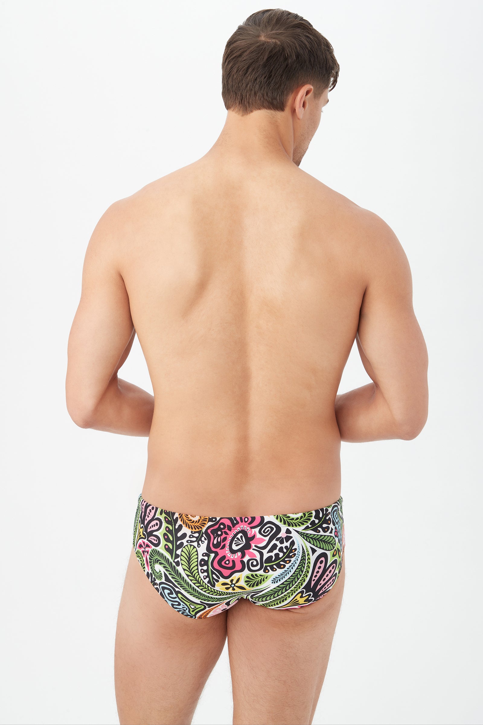 A man is seen from behind, standing shirtless and wearing Bondi Swim Briefs with colorful floral and abstract patterns on a white background.