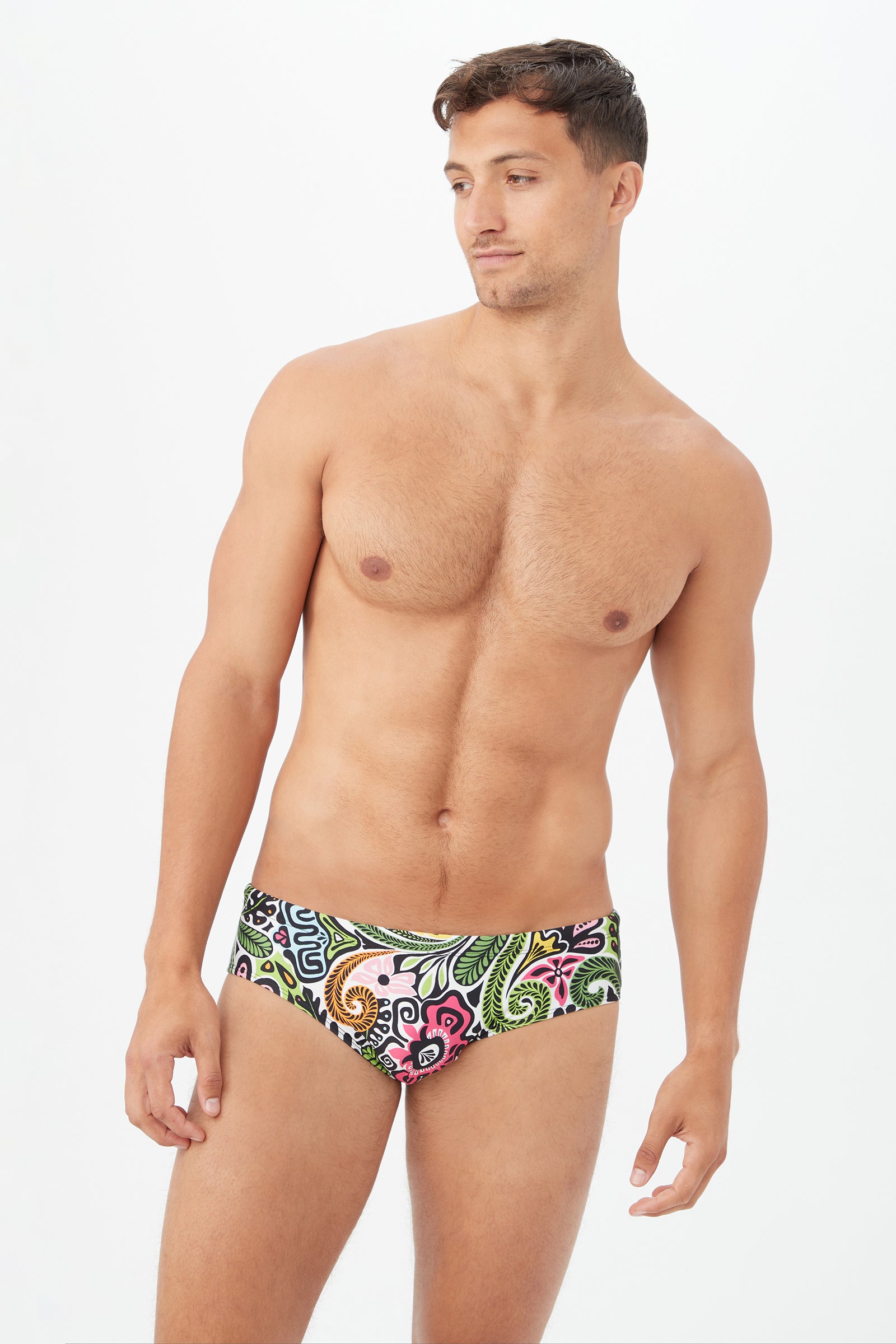 A man wears the Bondi Swim Brief in Multi, featuring a vibrant paisley pattern. He stands against a plain white background, looking slightly left with a relaxed expression and his right arm resting by his side.
