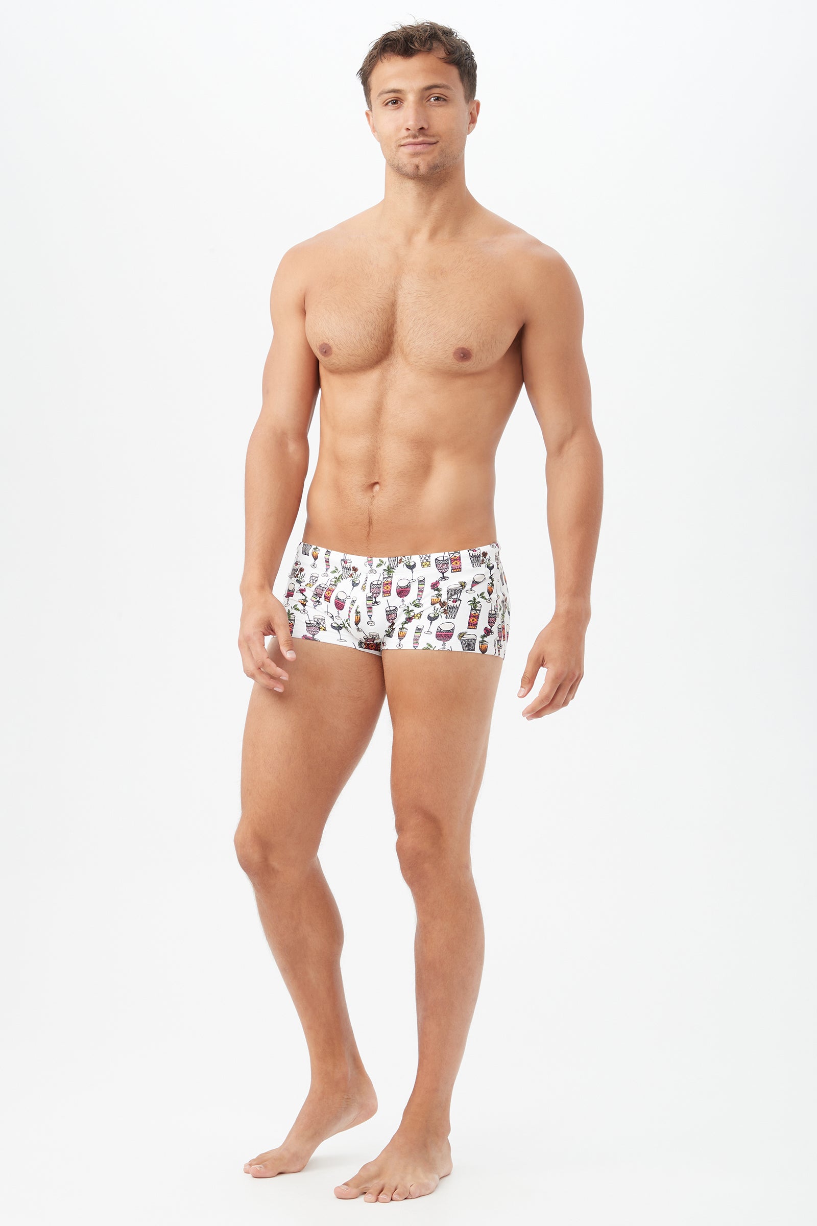 A man stands barefoot against a white background, facing forward with a neutral expression, wearing the Sorrento Swim Trunk – Happy Hour Swim Knit, featuring a vibrant and playful pattern.