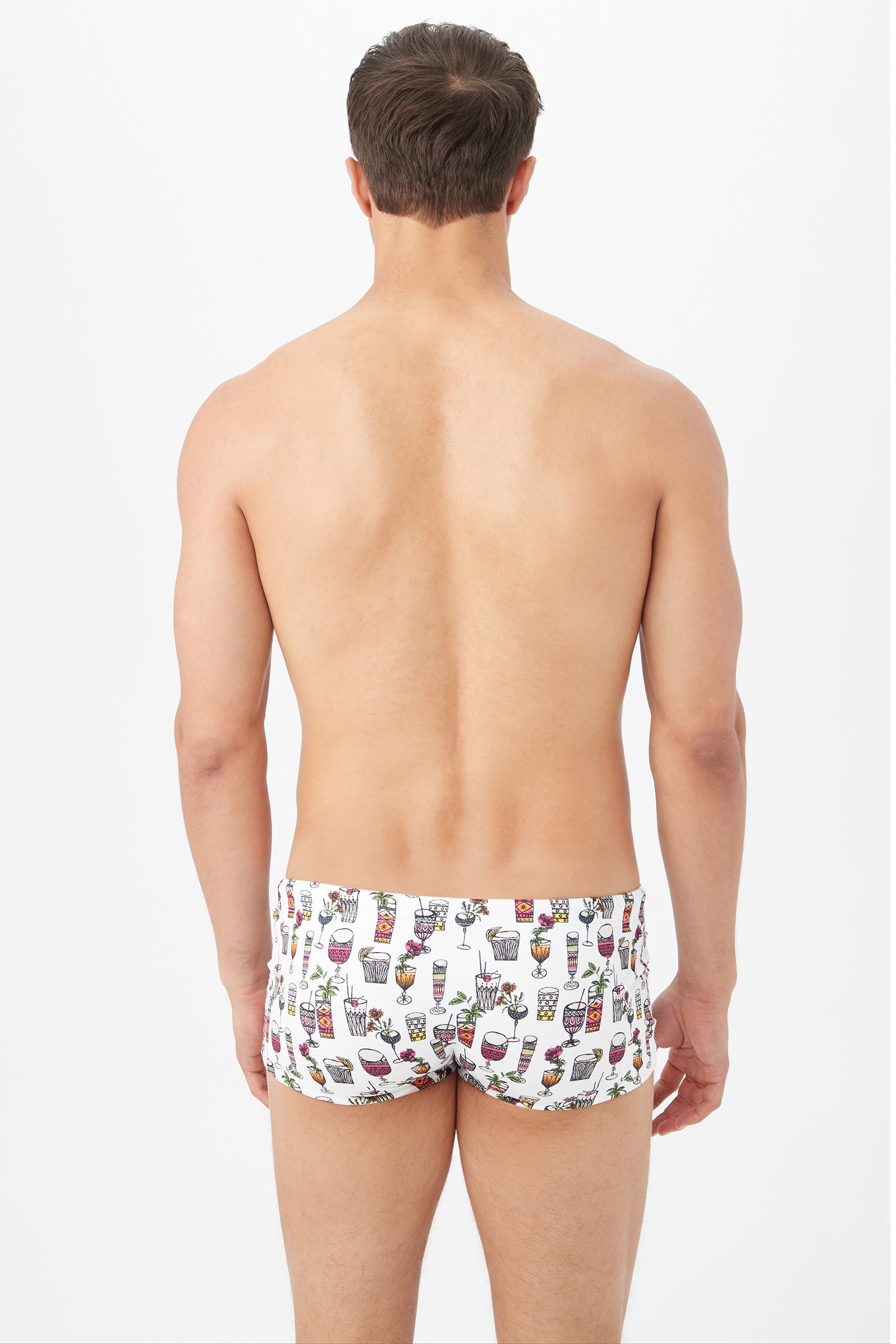 A man faces away from the camera, wearing the Sorrento Swim Trunk - Happy Hour Swim Knit, featuring a white base with colorful cocktail glass prints. The background is plain white.