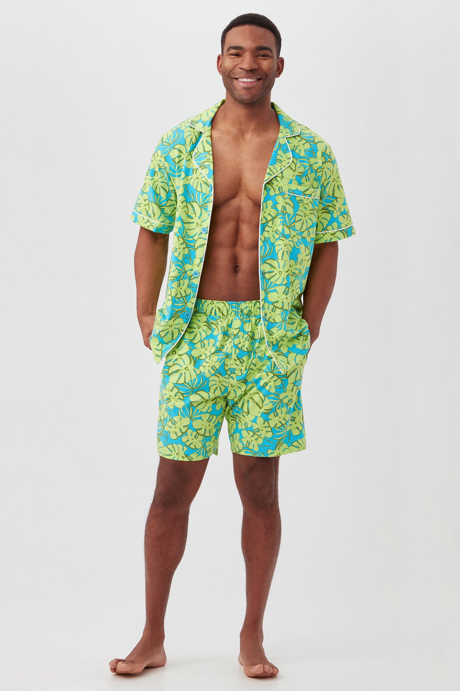 A smiling man stands barefoot in the Palm Leaves Mens Short Sleeve Boxer Short Jersey PJ Set—matching button-up shirt and shorts with a bright yellow and blue tropical floral pattern—posing against a plain white background.