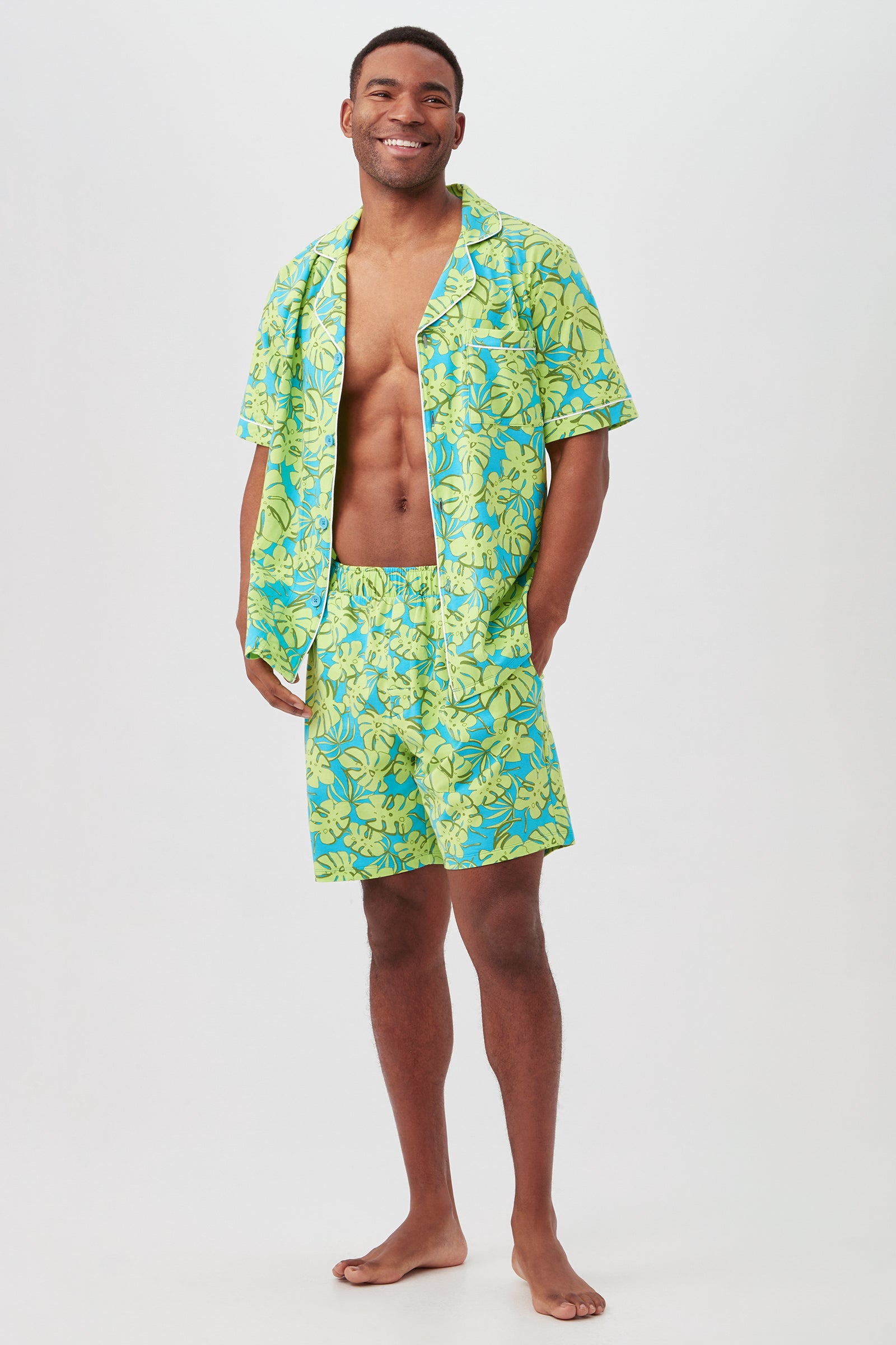 A man stands barefoot and smiling in the Palm Leaves Mens Short Sleeve Boxer Short Jersey PJ Set, featuring bright yellow and blue tropical leaves. The open shirt reveals his chest, making the vibrant set pop against a plain white background.