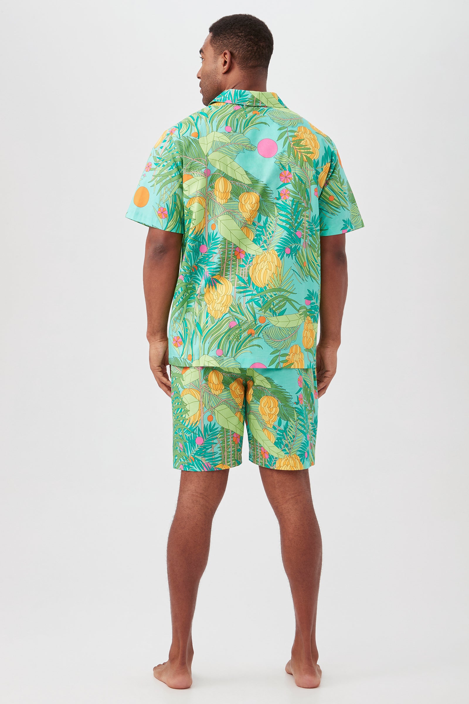 A man stands barefoot facing away, wearing the Going Bananas Mens Short Sleeve Boxer Short Poplin PJ Set—a stylish organic cotton pajama with a vibrant tropical pattern of green leaves, yellow pineapples, and pink accents on light blue.