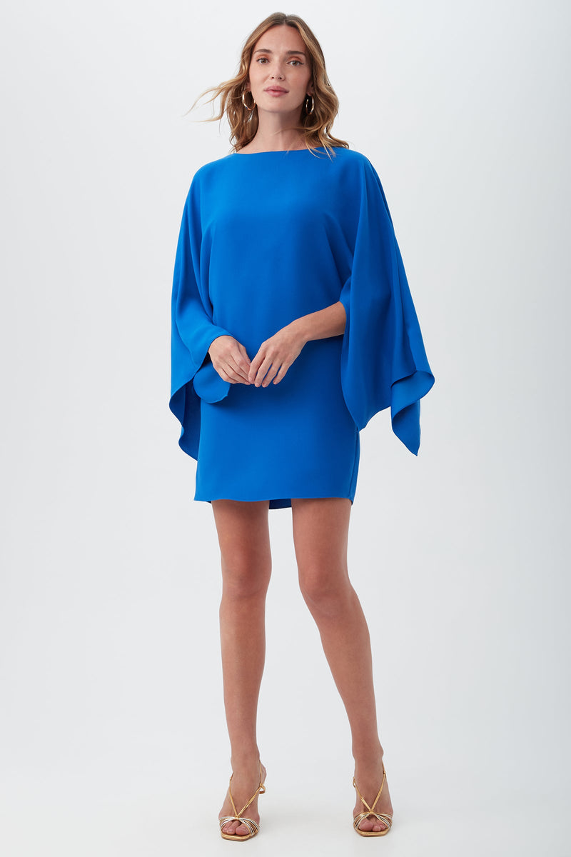A woman with light brown, wavy hair wears the Daiquiri Dress, a vibrant royal blue mini dress featuring wide batwing sleeves. She pairs it with gold open-toe heels and stands against a plain background.