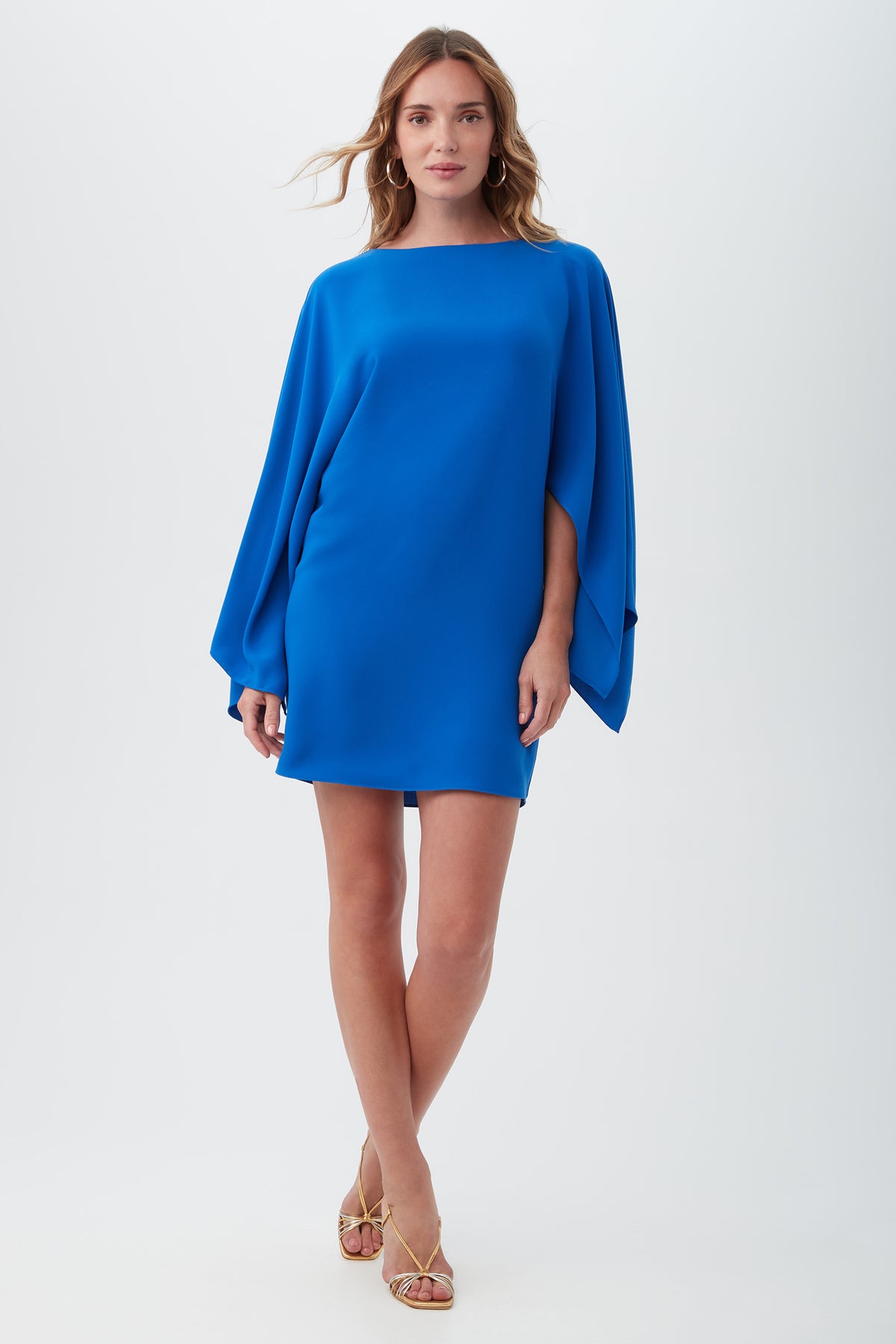 A woman wears the Daiquiri Dress, a loose-fitting bright blue mini dress with wide draped sleeves, paired with gold strappy heels against a plain white background.
