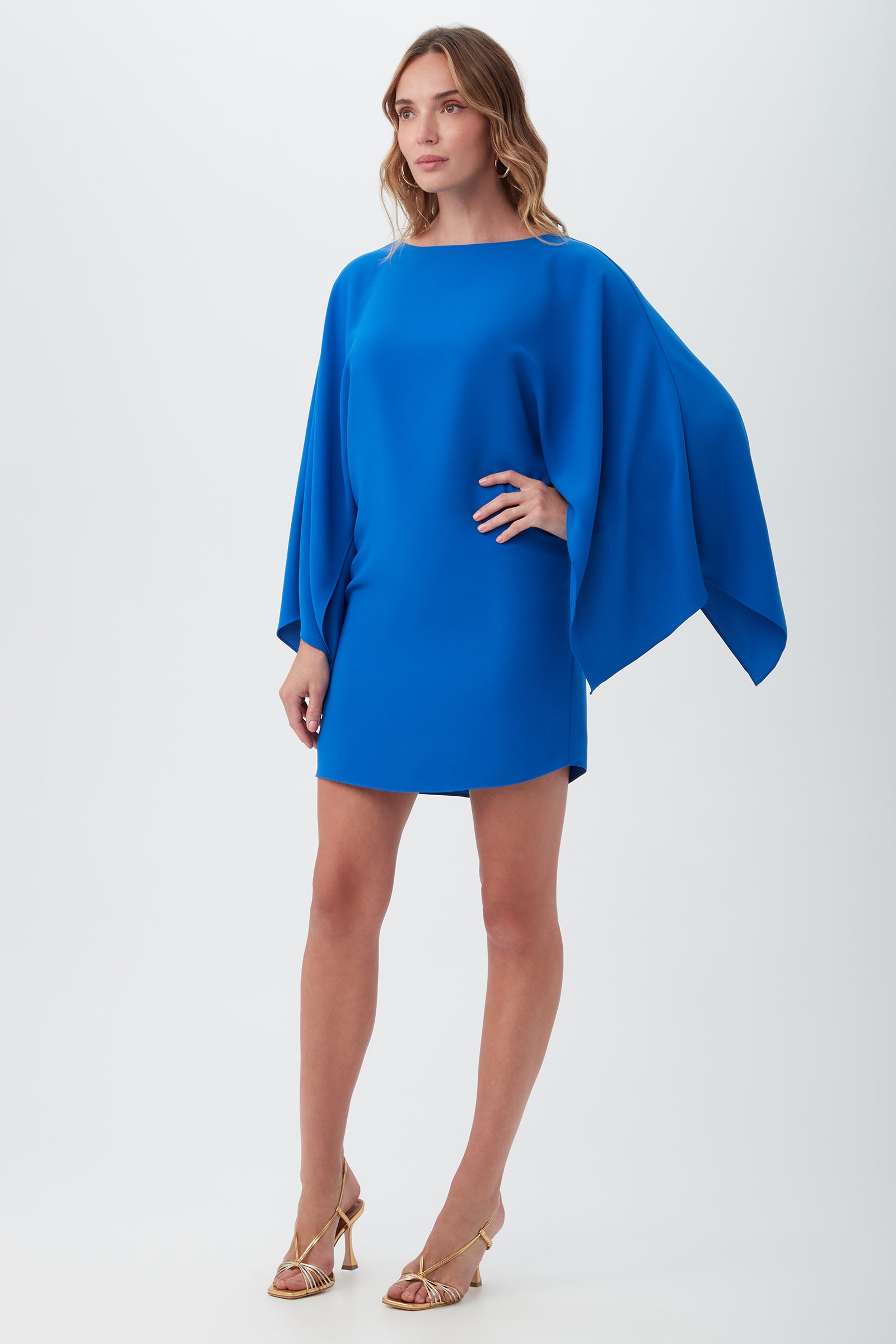 A woman poses in a studio against a plain light background, wearing the Daiquiri Dress—a vibrant blue, loose-fitting mini dress with wide, flowing sleeves—paired with gold strappy high-heeled sandals.