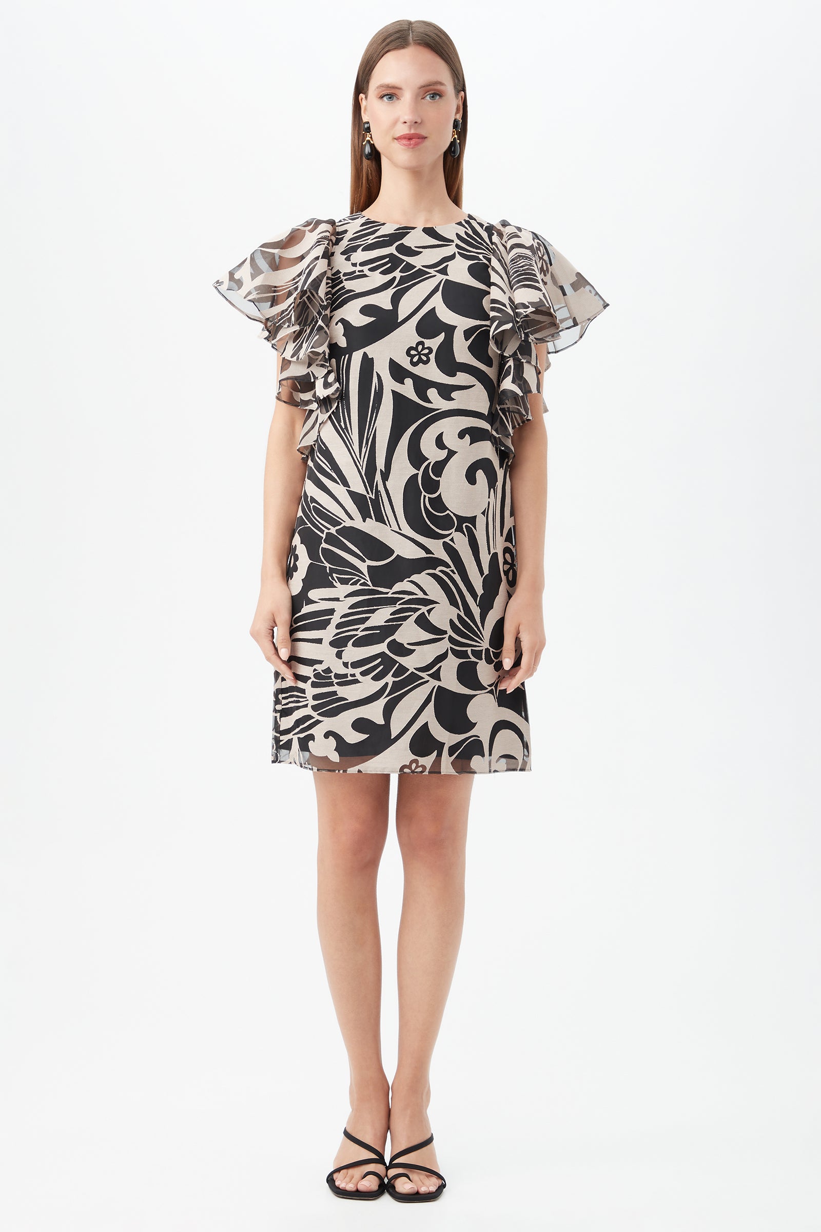 A woman faces forward wearing the Hokulani Dress, a short black and beige floral piece with sheer ruffled sleeves. She pairs it with black open-toe heels and minimal jewelry, standing against a plain white background.