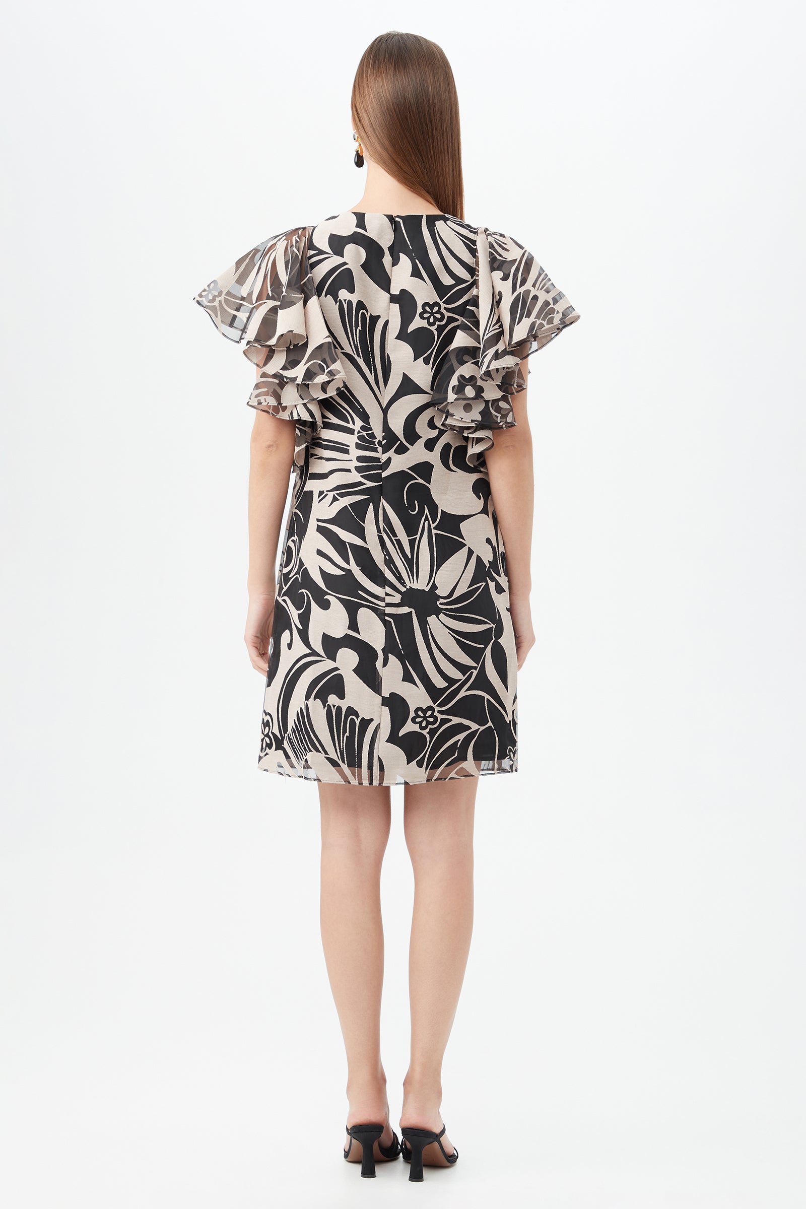 A woman faces away, wearing the Hokulani Dress—a short black-and-tan floral dress with flutter sleeves—and black heeled sandals. Her long, straight hair is loose, set against a plain white background.