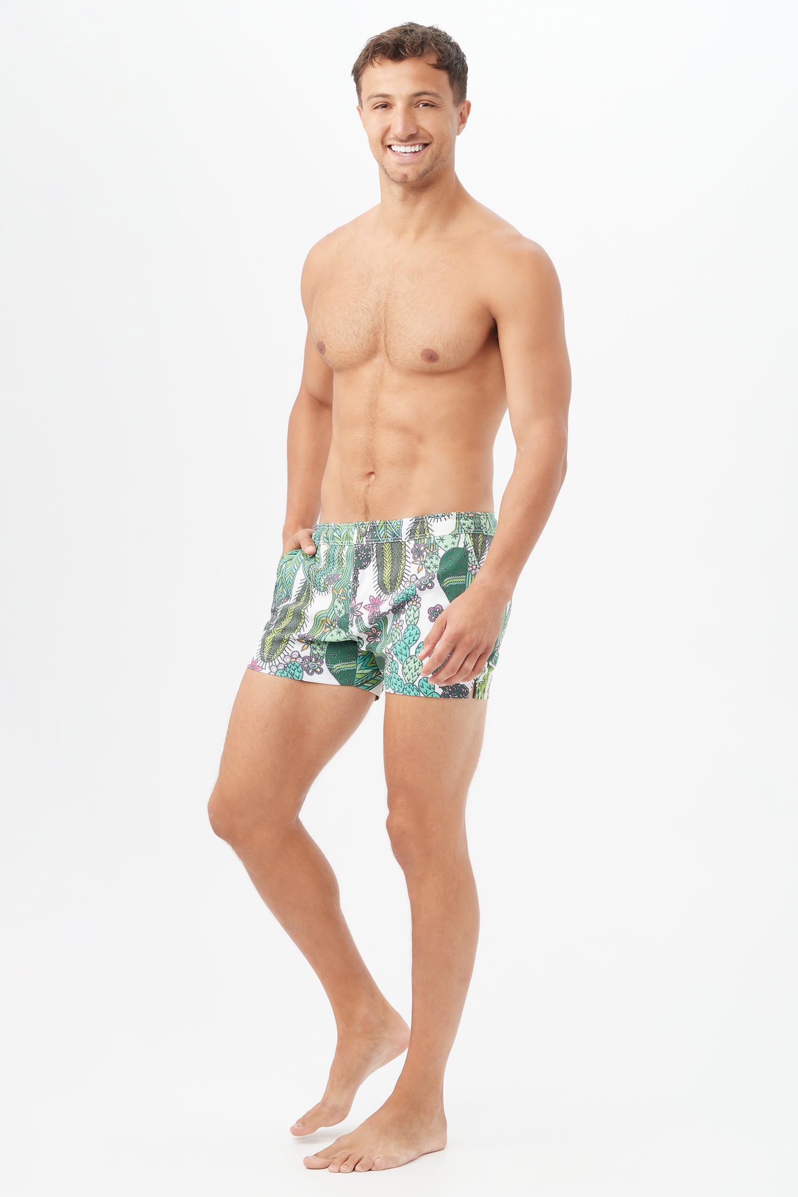 A smiling man stands barefoot against a white background, wearing the Chico Swim Trunk - Cacti Swim Woven. He has one hand in his pocket and a relaxed posture.