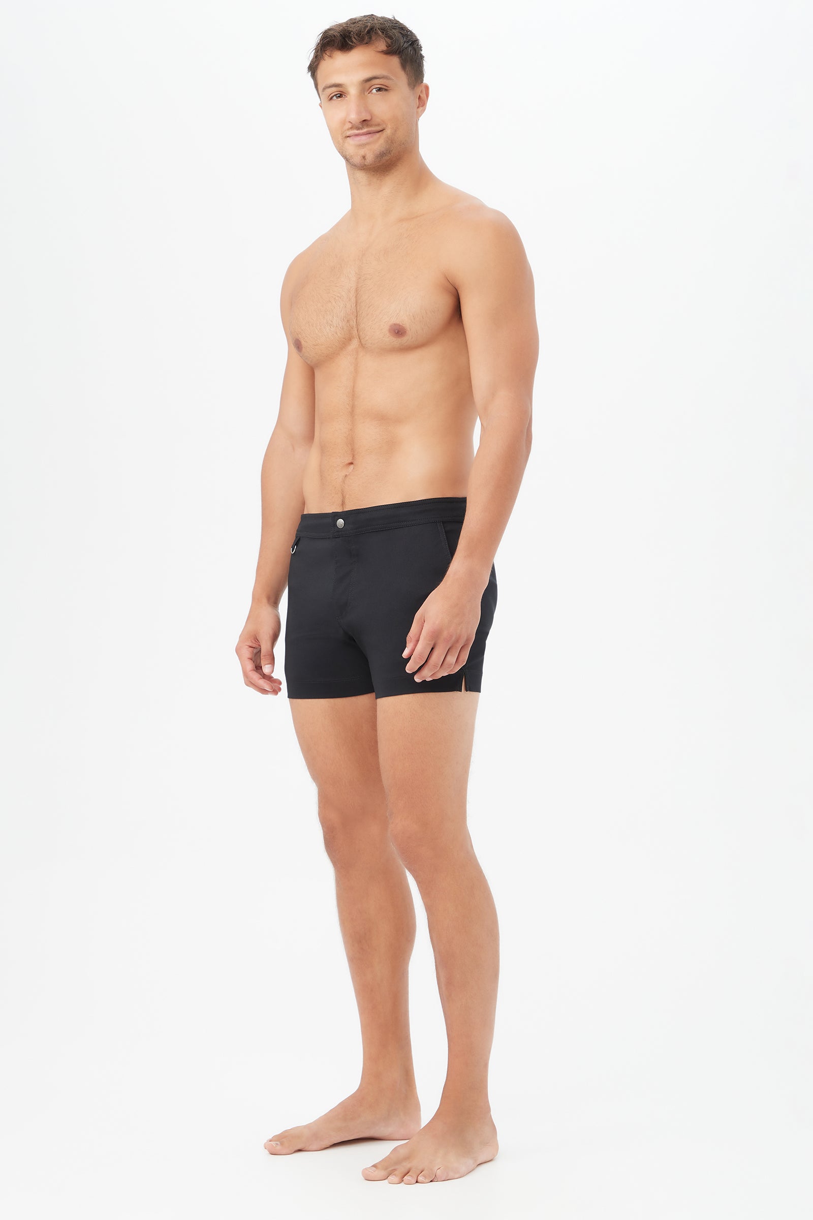A man with short brown hair stands barefoot against a white background, wearing Chico Swim Trunk - Metallic Swim Woven, smiling slightly while facing forward with his body angled to the side.