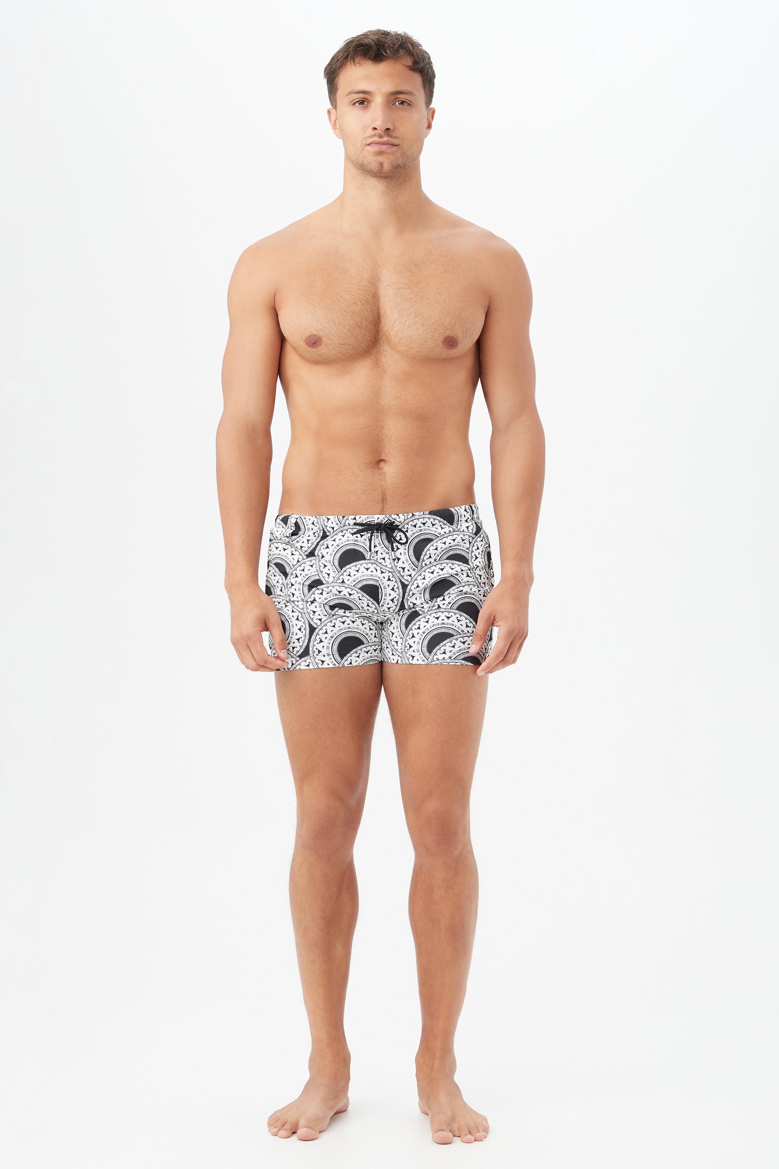 A man stands barefoot against a plain white background, wearing Palm Desert Swim Trunks in a black and white pattern. He faces forward with a neutral expression and his arms relaxed at his sides.