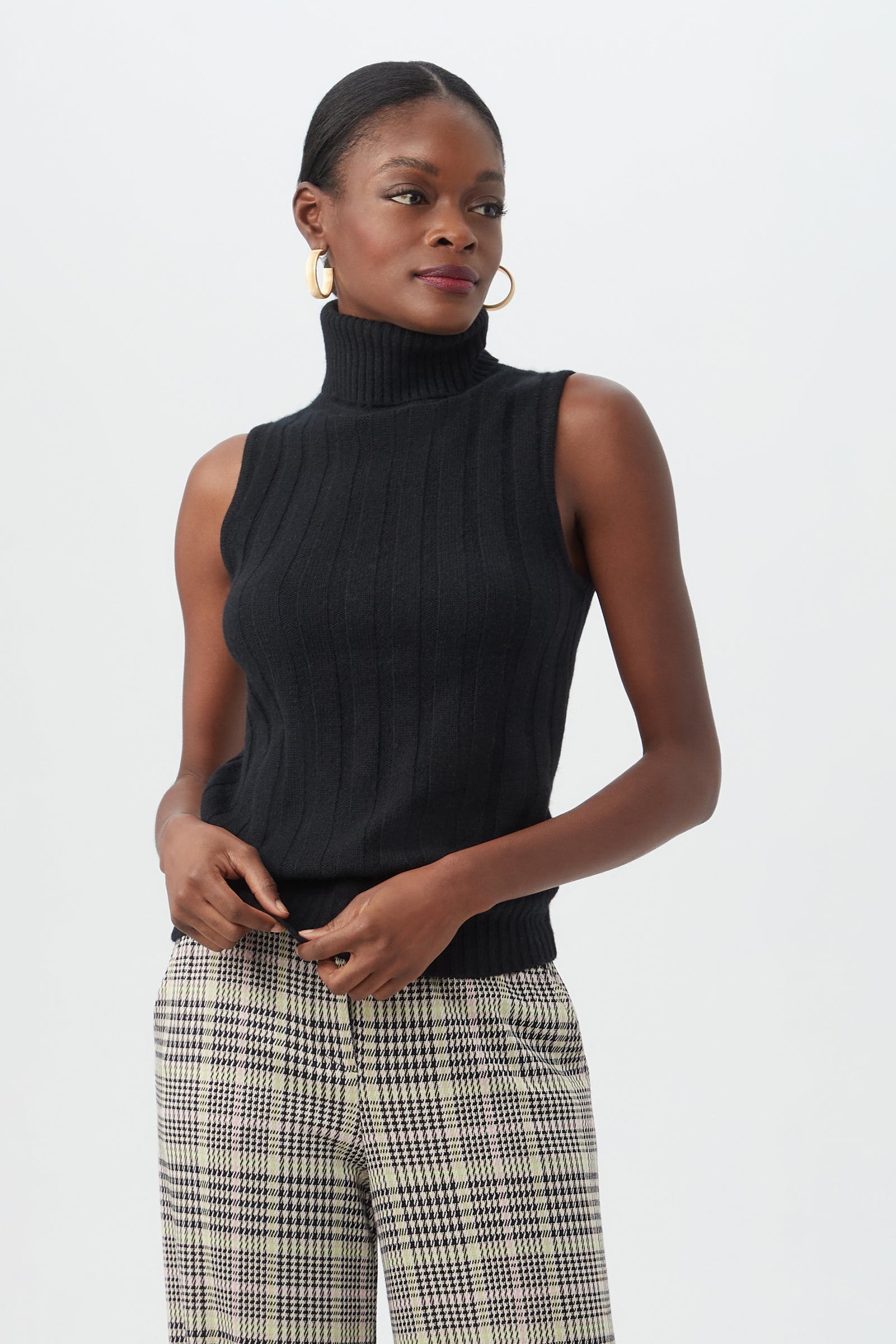 Wearing the Onassis Knit Top, a woman with her hair pulled back and gold hoop earrings stands against a white background in plaid trousers, looking slightly to the side.
