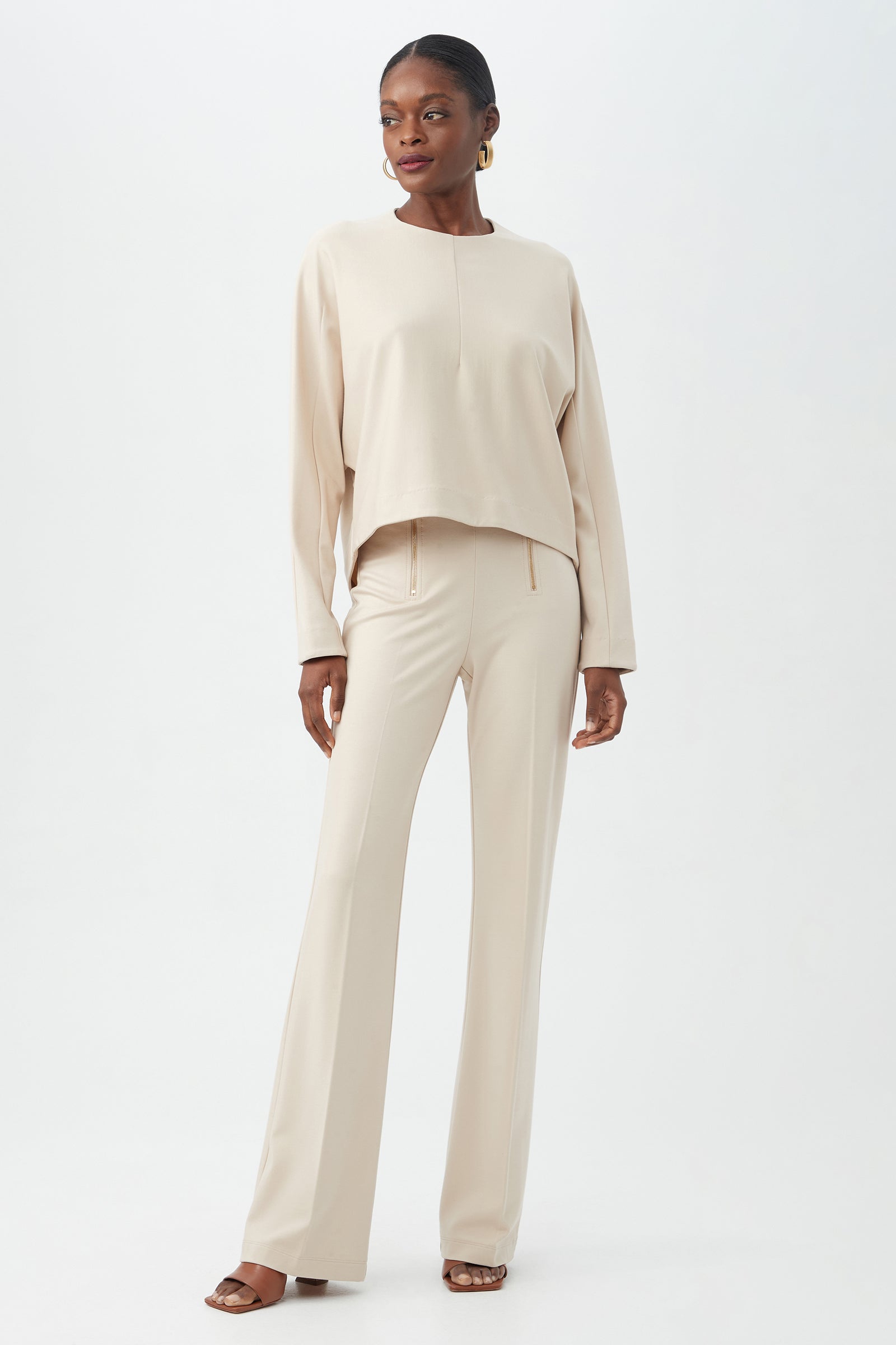 A woman poses against a plain background wearing the Kiara Pant with a loose, long-sleeve beige top, gold hoop earrings, and brown heeled sandals.