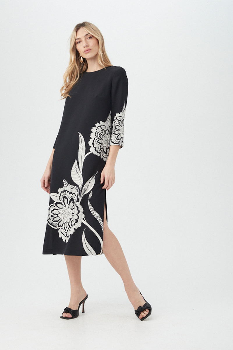 A woman models the Cointreau 2 Dress in Black/Savanna Sand, featuring a mid-length cut, three-quarter sleeves, a side slit, and contrasting floral patterns. She pairs it with black heels. Her long wavy blonde hair stands out against a white background.
