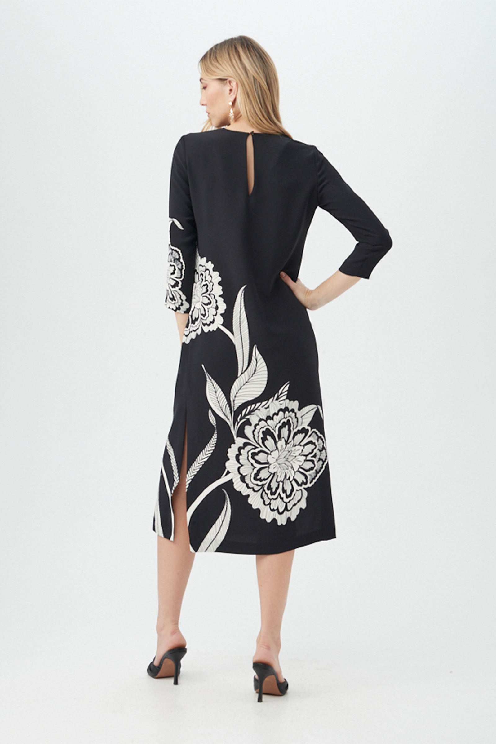 A woman faces away wearing the Cointreau 2 Dress—a black, knee-length piece with white floral prints, three-quarter sleeves, back keyhole and slits—paired with black heeled sandals against a plain white background.