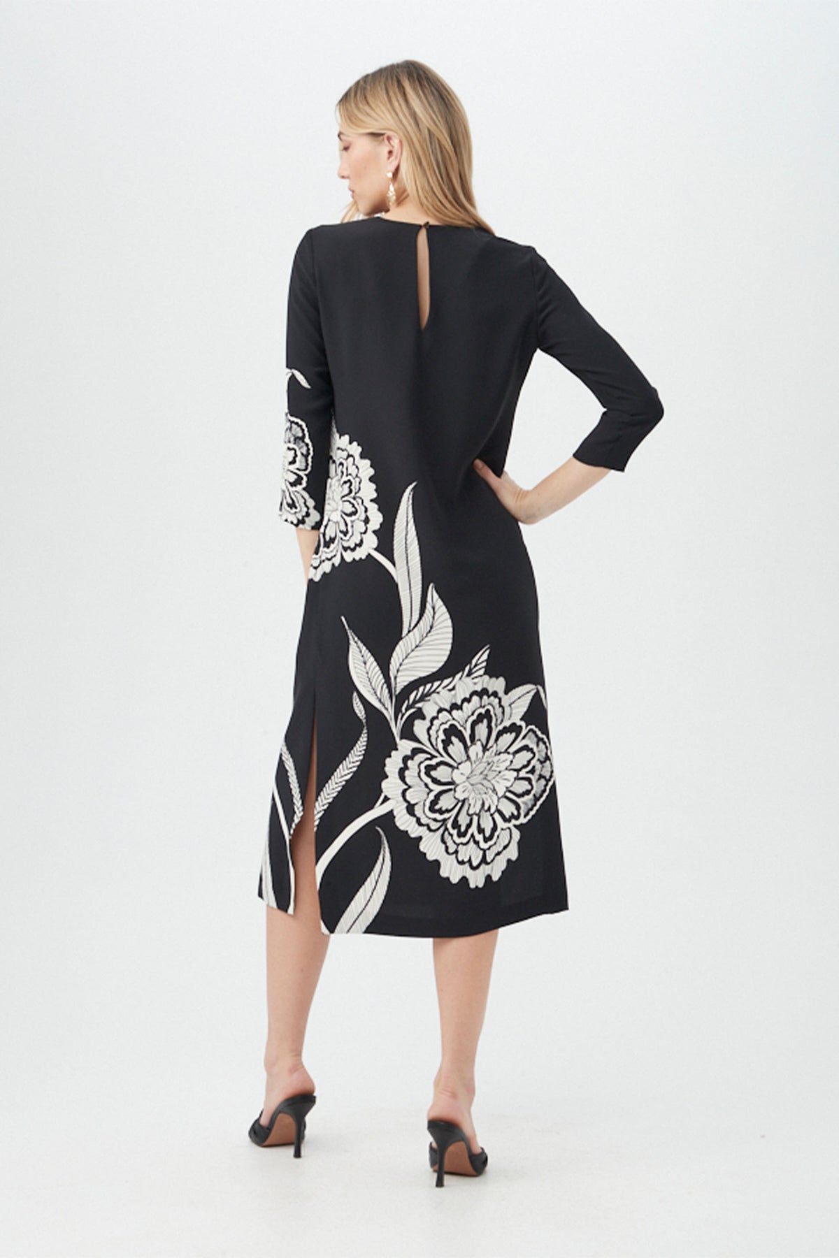 A woman faces away wearing the Cointreau 2 Dress—a black, knee-length piece with white floral prints, three-quarter sleeves, back keyhole and slits—paired with black heeled sandals against a plain white background.