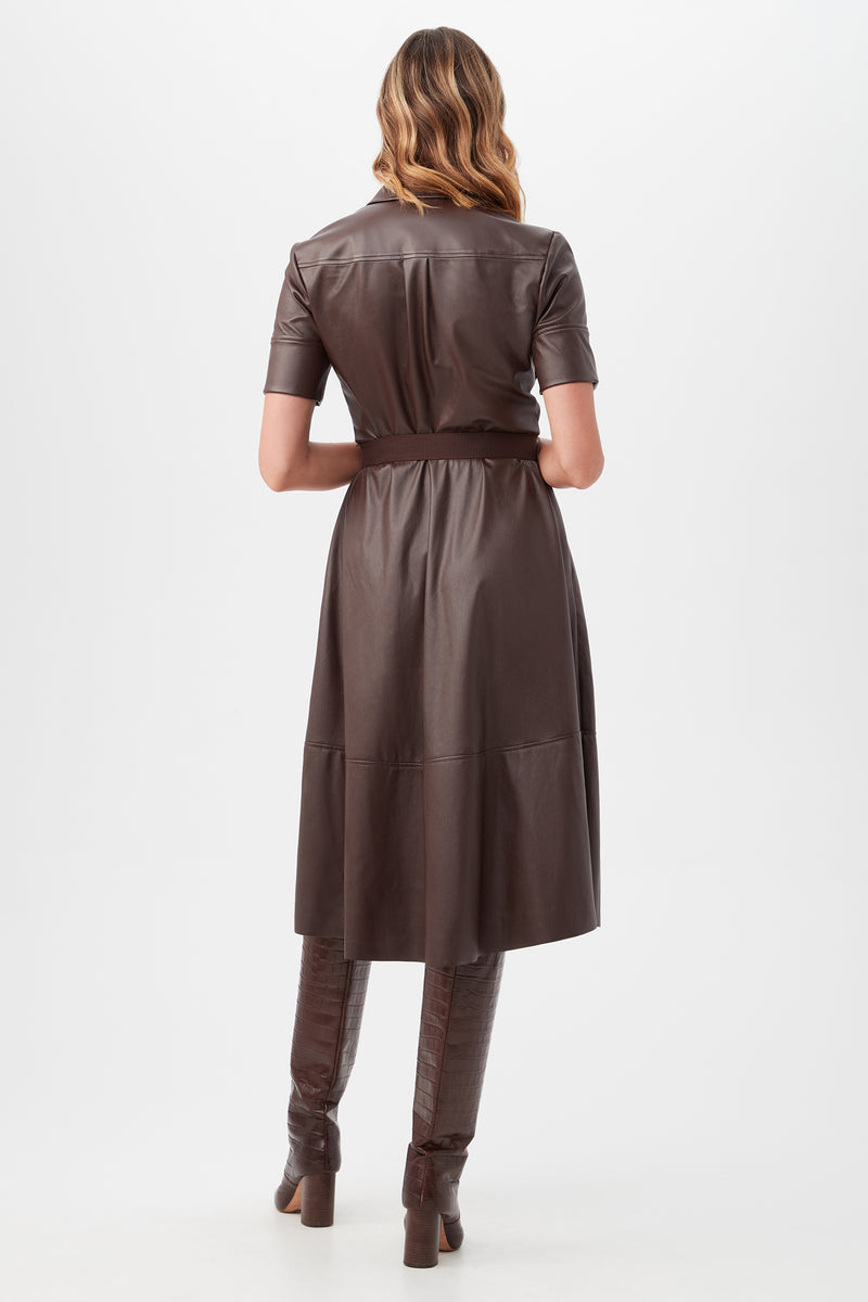 A woman with wavy hair faces away, wearing the brown, short-sleeved, belted Misola Dress with matching tall heeled boots, standing against a plain white background.