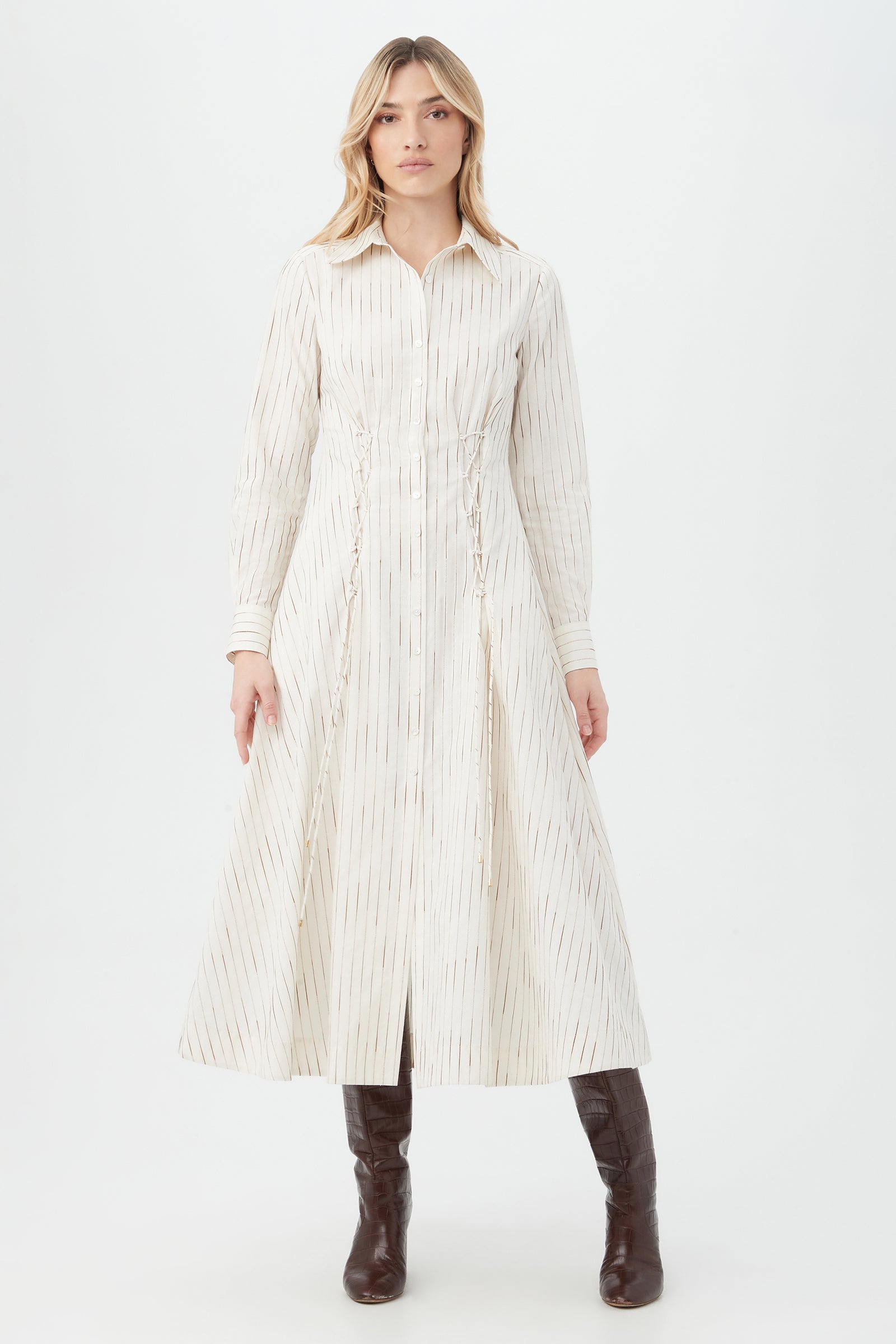A woman stands against a white background wearing the Omari Dress in White/Camel, a long-sleeved midi with thin vertical stripes, paired with brown knee-high boots. She faces forward with a neutral expression.