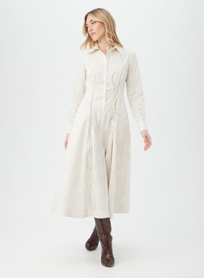 A woman wears the Omari Dress—a cream, long-sleeve, collared midi dress with subtle vertical stripes—paired with dark brown knee-high boots, standing against a plain white background.
