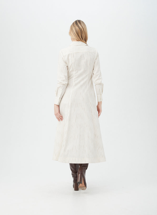 A woman with blonde hair stands facing away, wearing the long-sleeved, ankle-length Omari Dress in cream with subtle vertical stripes and knee-high brown boots against a plain white background.