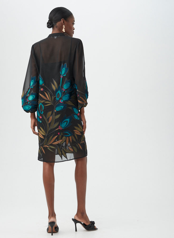 A woman faces away, wearing the Boheme 2 Dress—a sheer black dress with vibrant floral and leaf patterns on the back. Her hair is styled in a low bun, paired with black heeled sandals.