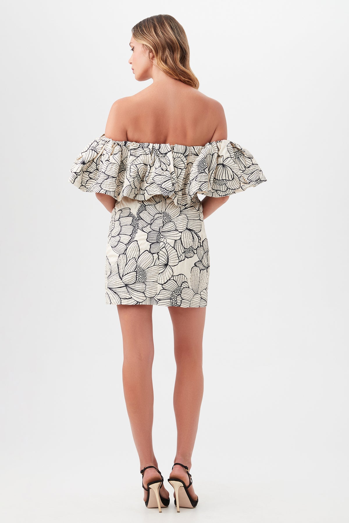 A woman faces away, wearing the Nia Dress—an off-the-shoulder, ruffled mini dress with a bold black and white floral print—paired with high-heeled sandals against a plain white background.