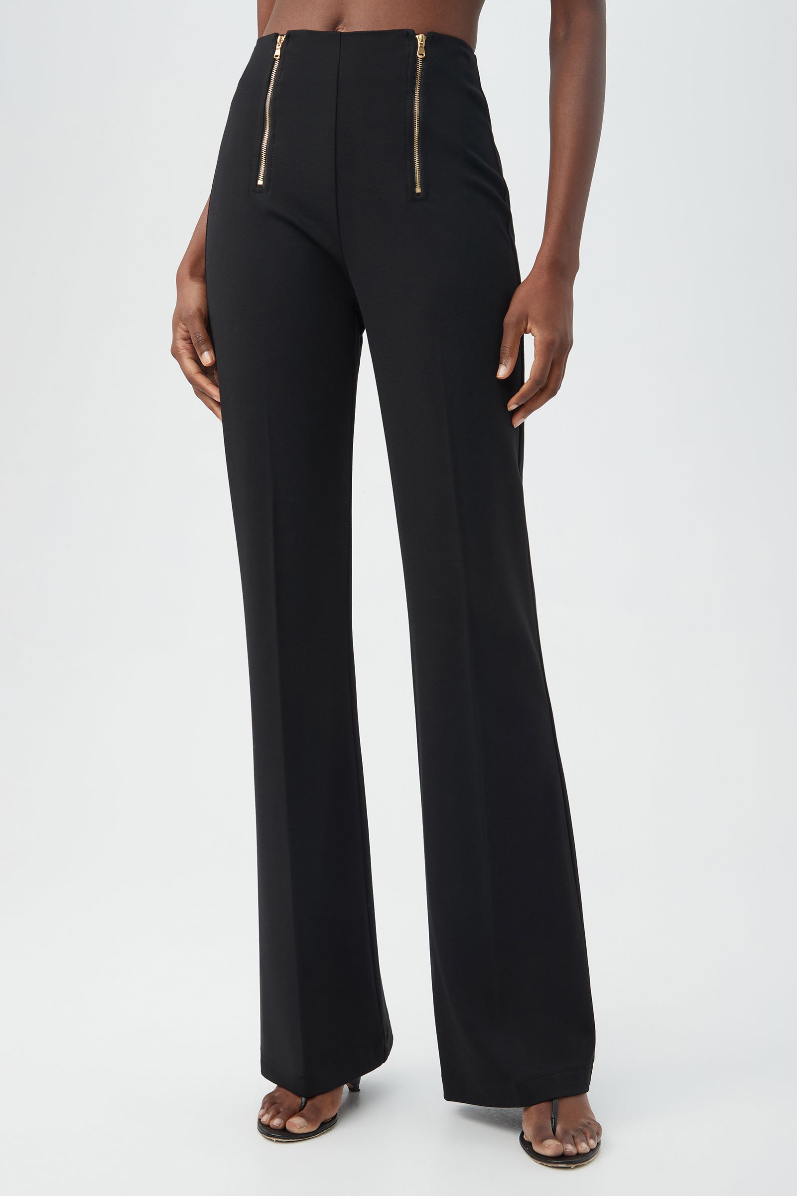 A person wears the Kiara Pant in black—high-waisted, wide-leg style with two gold front zippers—paired with open-toe heels against a plain light background.