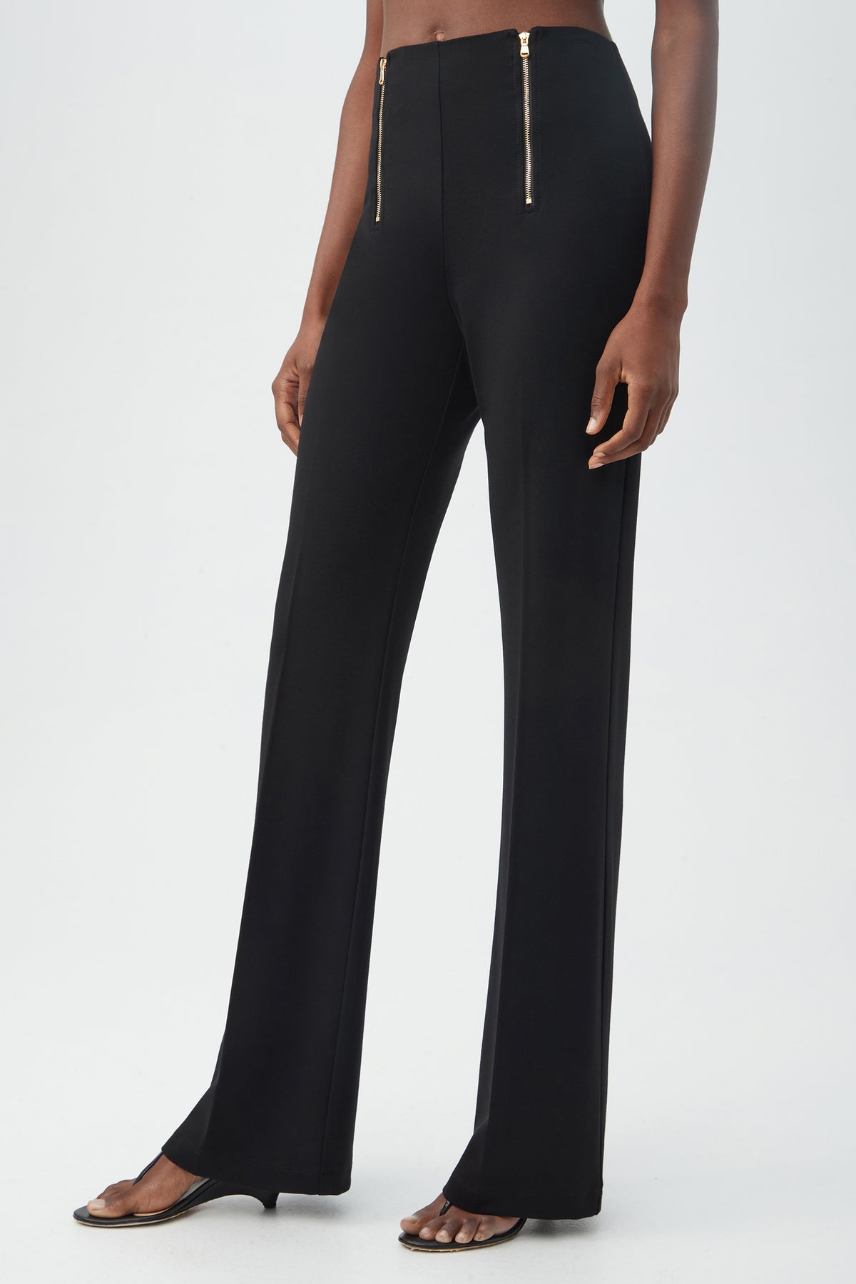 A person wearing the Kiara Pant—high-waisted, black, wide-leg pants with two gold front zippers—paired with open-toe heeled sandals, standing against a plain background.