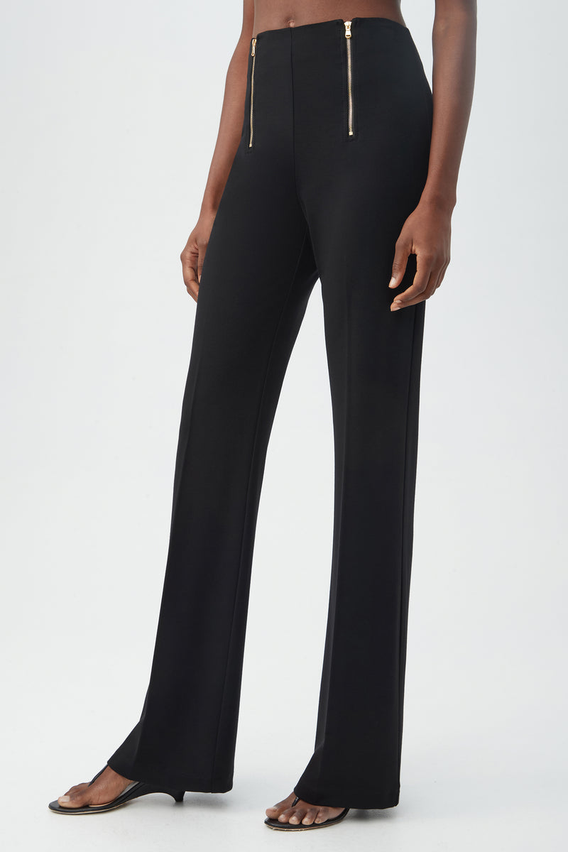 A person wearing the Kiara Pant—high-waisted, black, wide-leg pants with two gold front zippers—paired with open-toe heeled sandals, standing against a plain background.