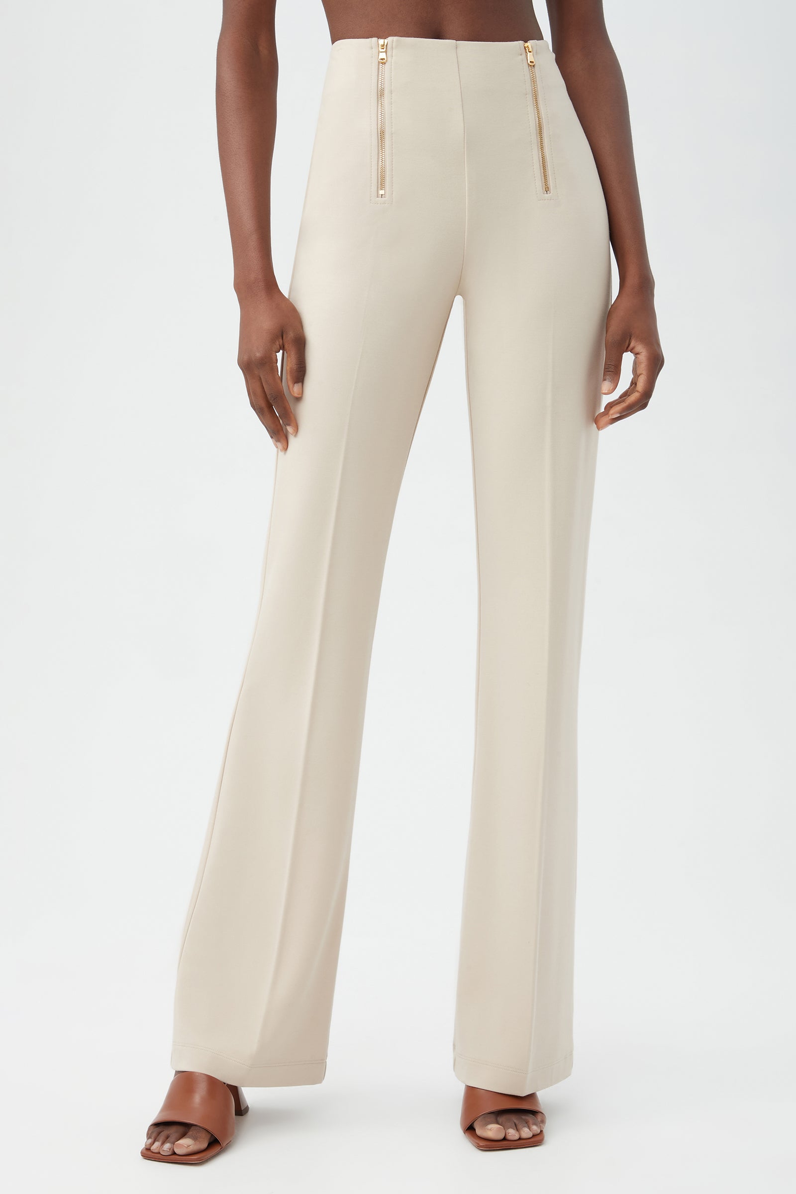A woman models the Kiara Pant in Sahara Stone, featuring a high waist, wide-leg silhouette, and gold hip zippers. She stands in brown heeled sandals against a plain white background.