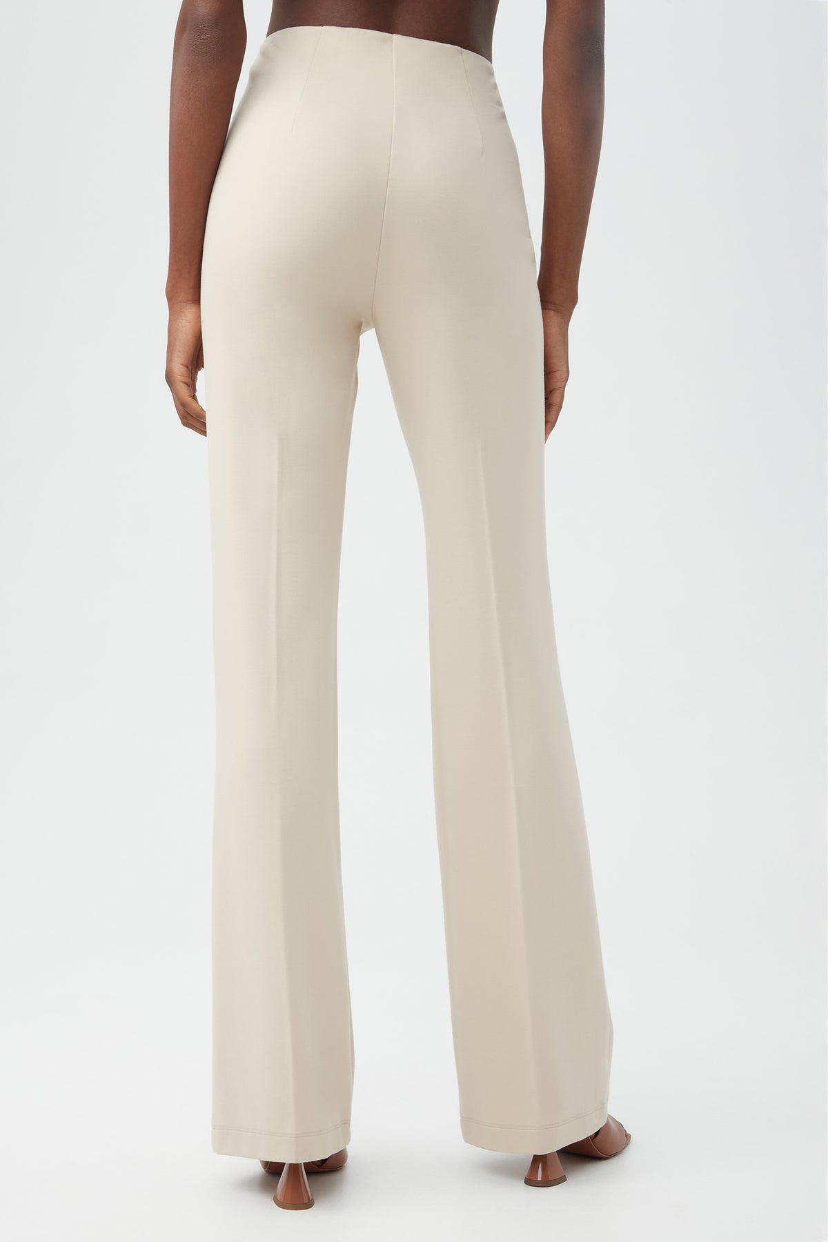 A person stands facing away, wearing the Kiara Pant—high-waisted, light beige, wide-leg trousers—and open-toe heels against a plain white background.