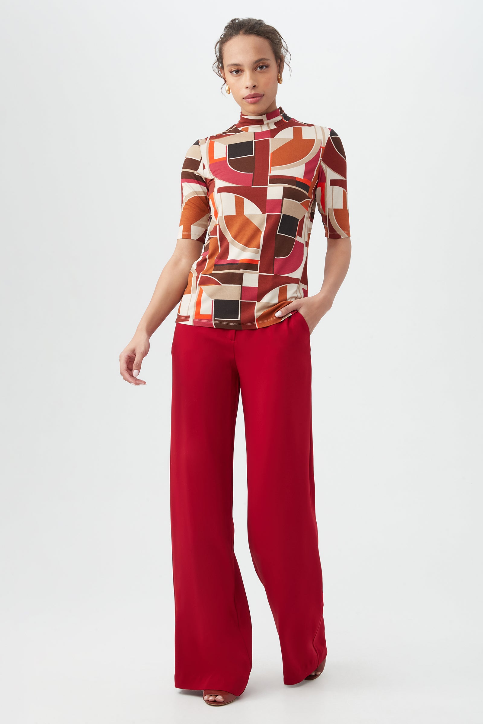 A person stands against a plain background wearing the Pierce 3 Topâ€â€a geometric patterned earth-tone topâ€â€with high-waisted, wide-leg red pants and one hand in their pocket.