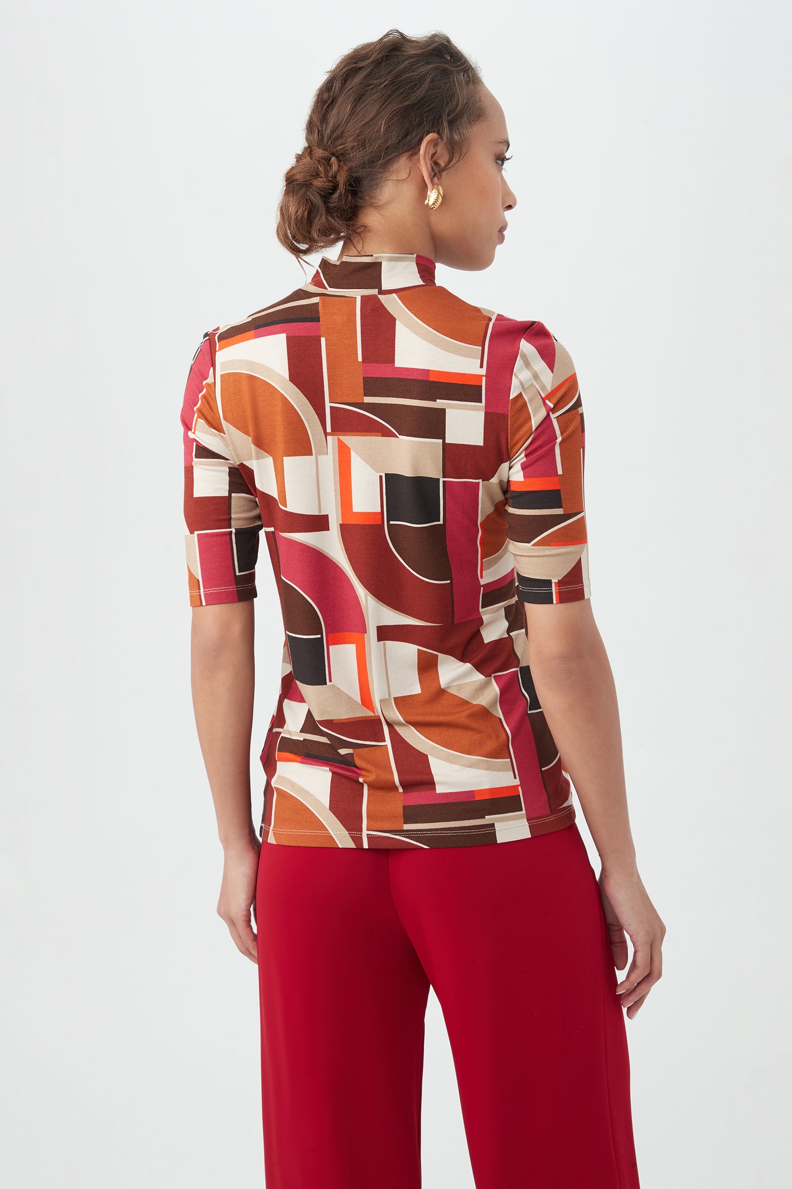 A woman with braided hair faces away, wearing the Pierce 3 Topâ€â€a geometric-patterned top in brown, red, orange, and creamâ€â€paired with solid red pants against a plain white background.