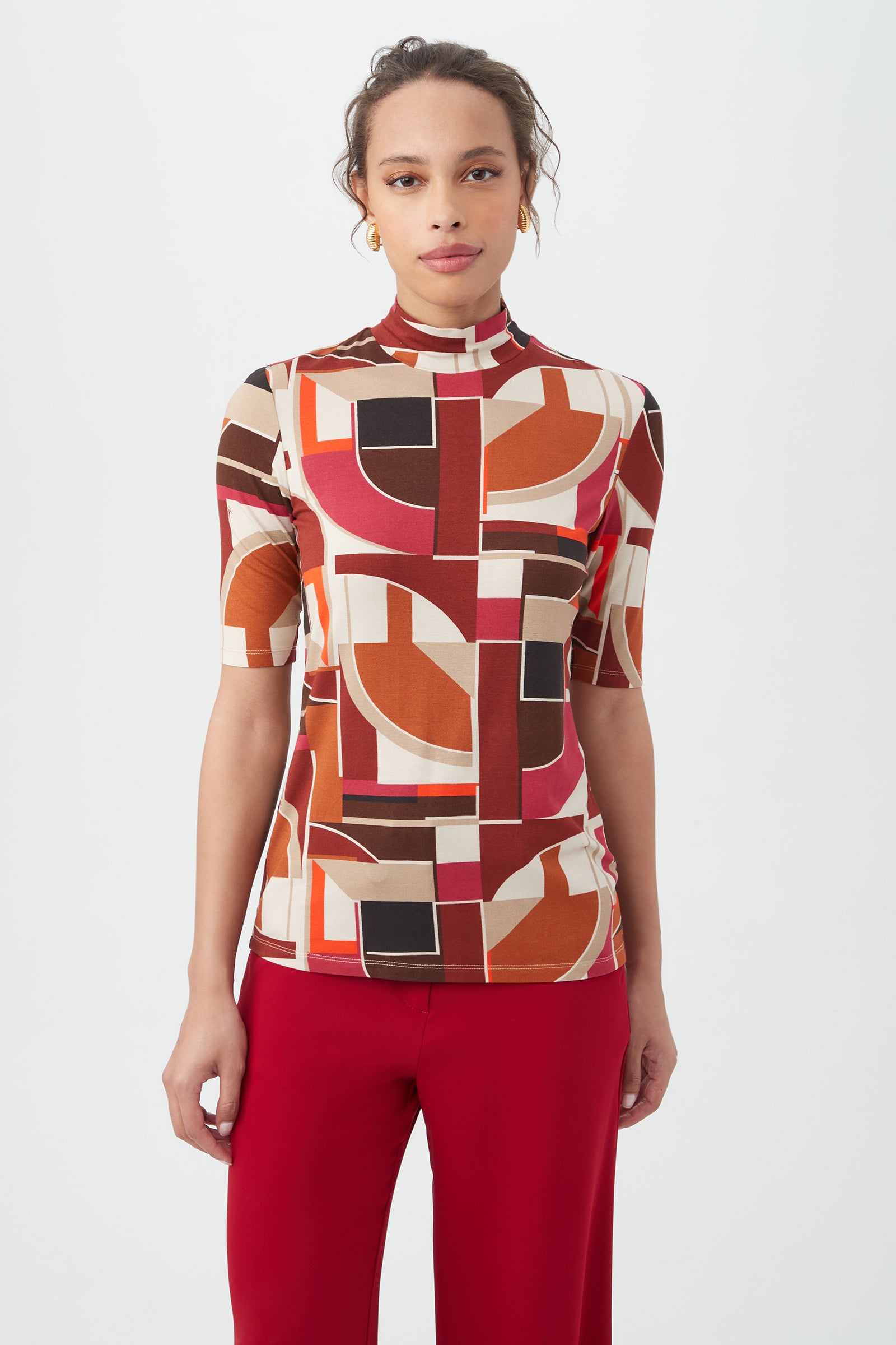 A woman stands against a white background wearing the Pierce 3 Top in Multi, featuring a colorful geometric pattern with short sleeves and abstract shapes in red, orange, beige, and black, paired with red pants.
