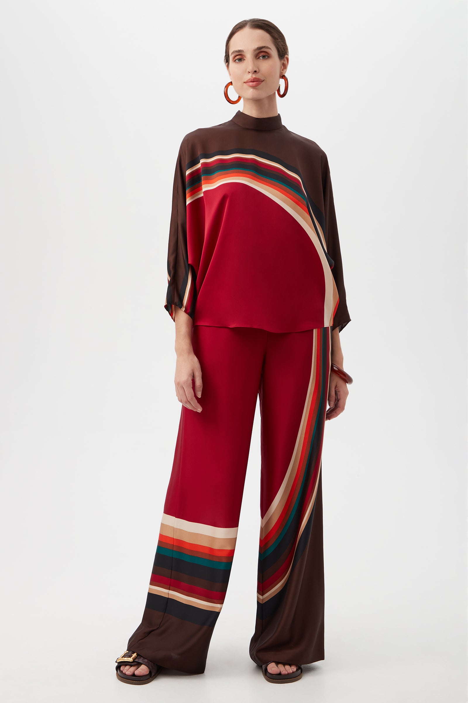 A woman stands against a plain background wearing the loose-fitting Kobina Top, featuring bold horizontal and diagonal stripes in red, brown, green, and beige. She accessorizes with hoop earrings and slip-on sandals.