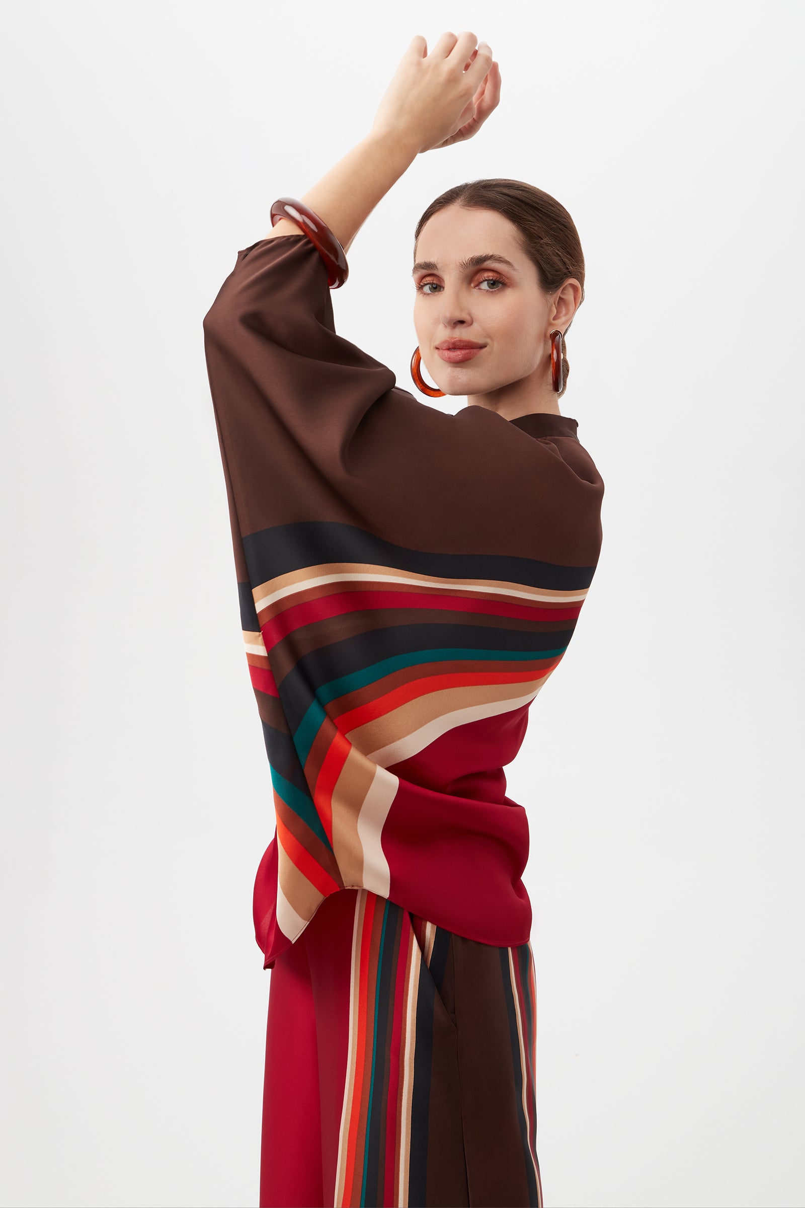 A woman confidently poses against a white background, raising one arm while wearing the Kobina Top—a vibrant piece featuring bold horizontal stripes in red, brown, green, and beige.